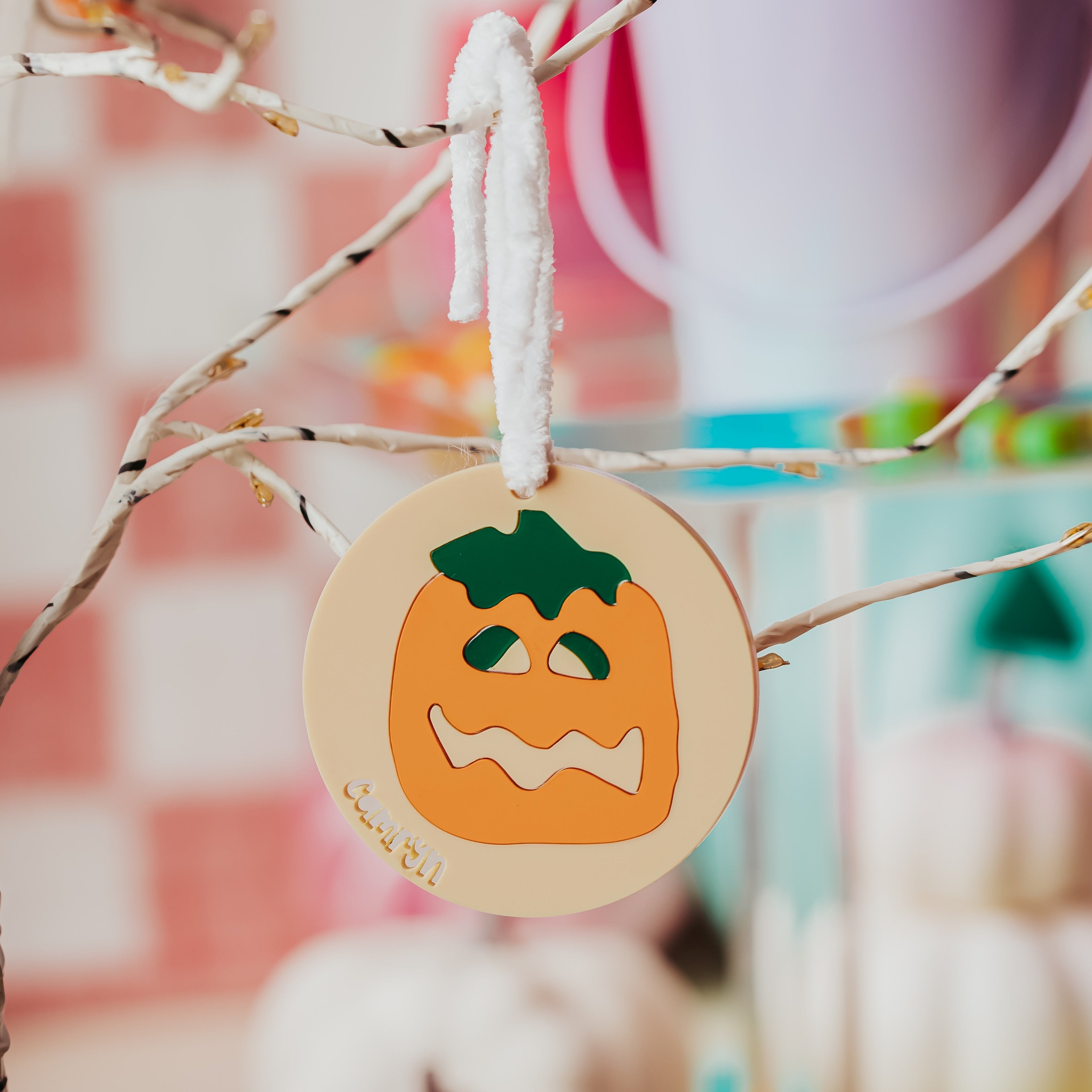 A round acrylic Halloween sugar cookie bag tag featuring a carved orange pumpkin face with a green stem, hanging from a white string on a decorative branch in a softly blurred fall-themed setting.
