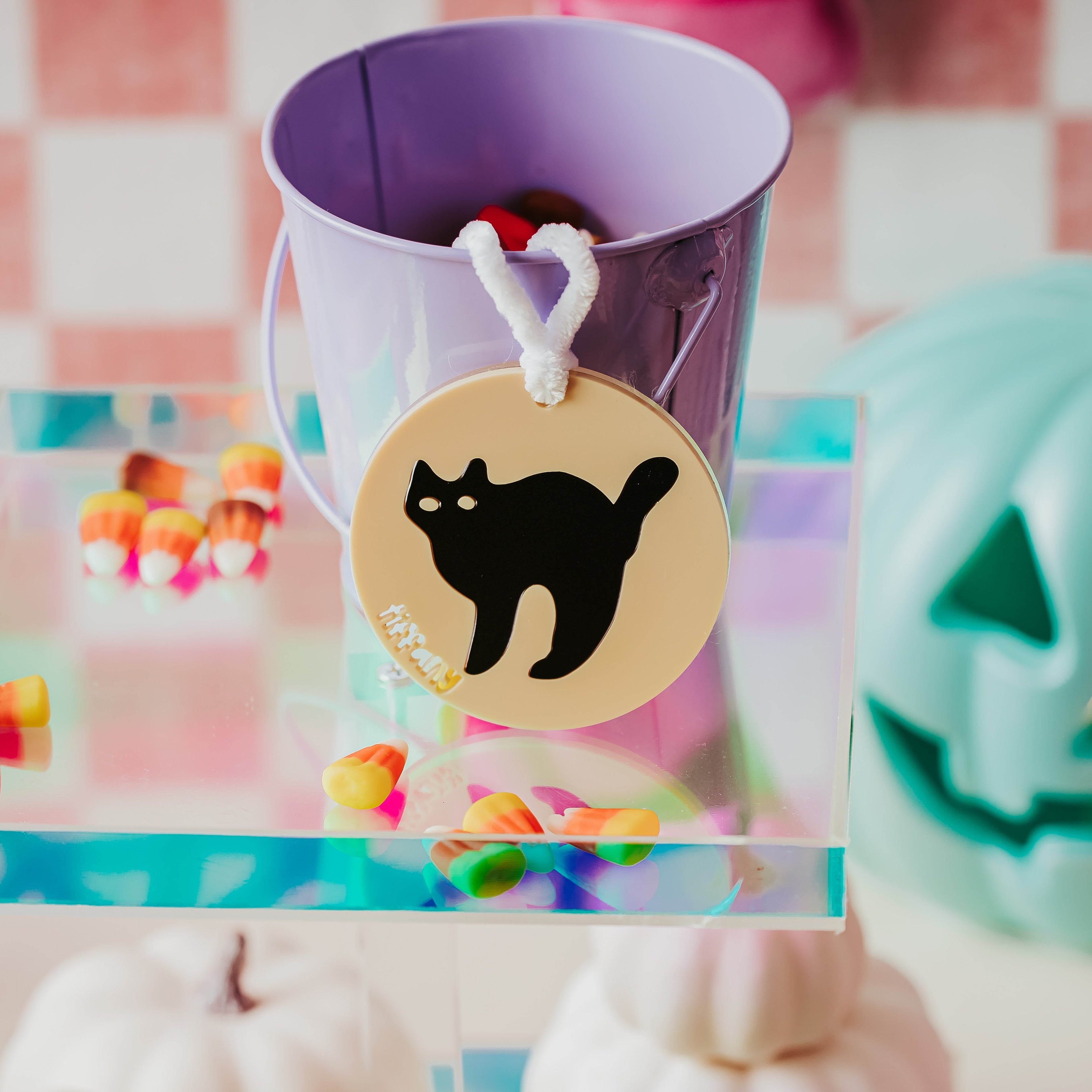 A acrylic Halloween sugar cookie basket tag display featuring a lavender bucket filled with candy, adorned with a round beige tag printed with a black cat silhouette and the word 'tiffany'. The bucket sits on a clear acrylic stand decorated with candy, with a teal jack-o'-lantern and pink checkered background visible.