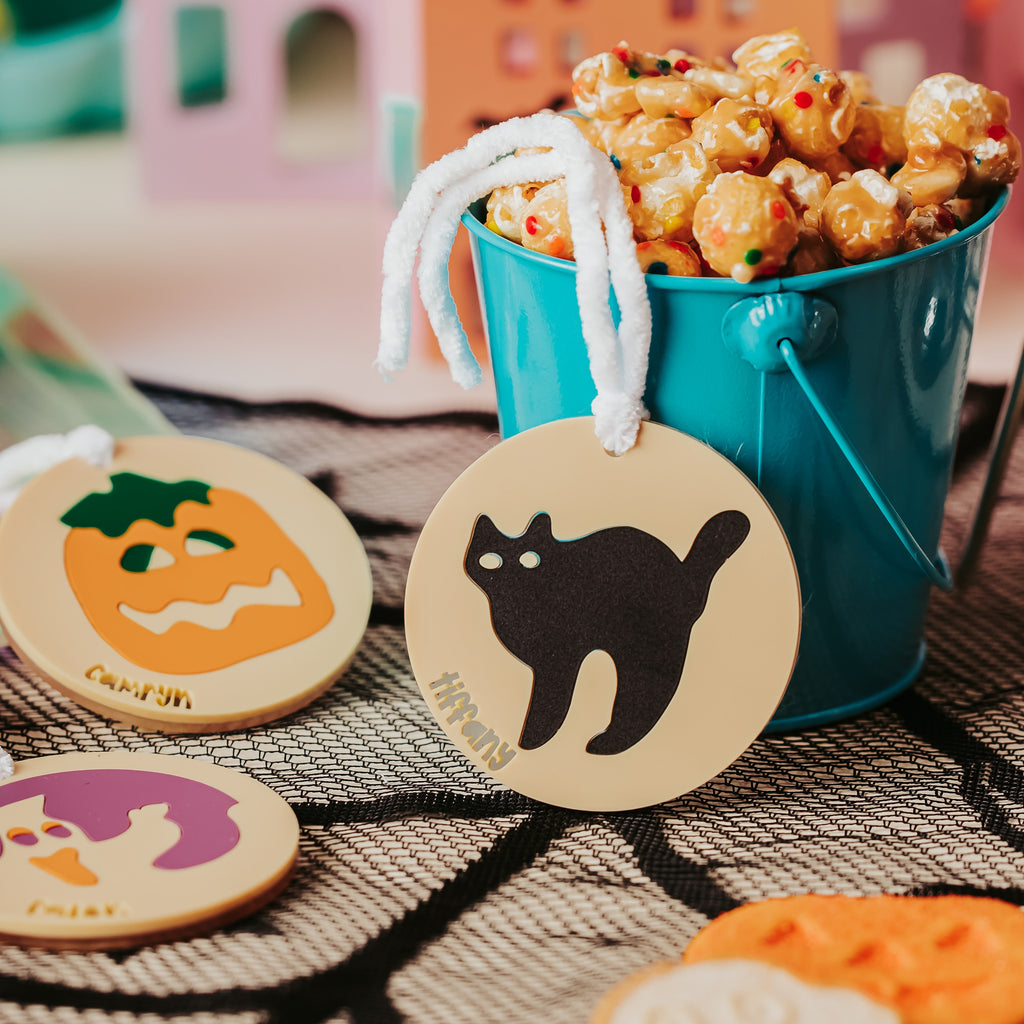 An acrylic Halloween sugar cookie basket tag with snack setup with a turquoise bucket filled with popcorn and candy in the background