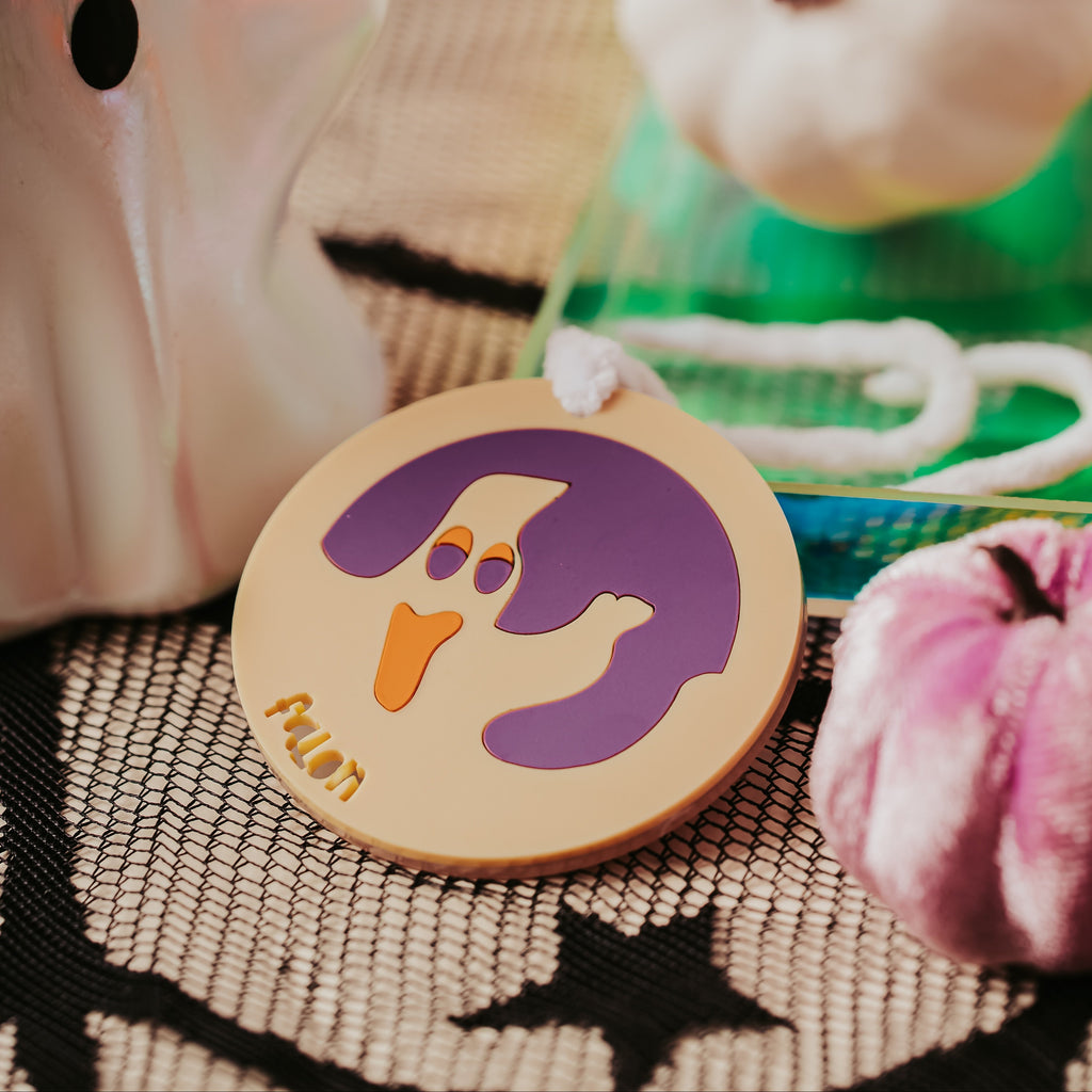 A close-up of an halloween themed acrylic sugar cookie bag tag featuring a purple ghost design with a small orange face and the name 'falon' cut out from beige portion. The ornament is styled on a black-and-white patterned fabric with Halloween-themed props, including a white ghost-shaped candle, a white pumpkin, and a velvet pink decorative pumpkin.