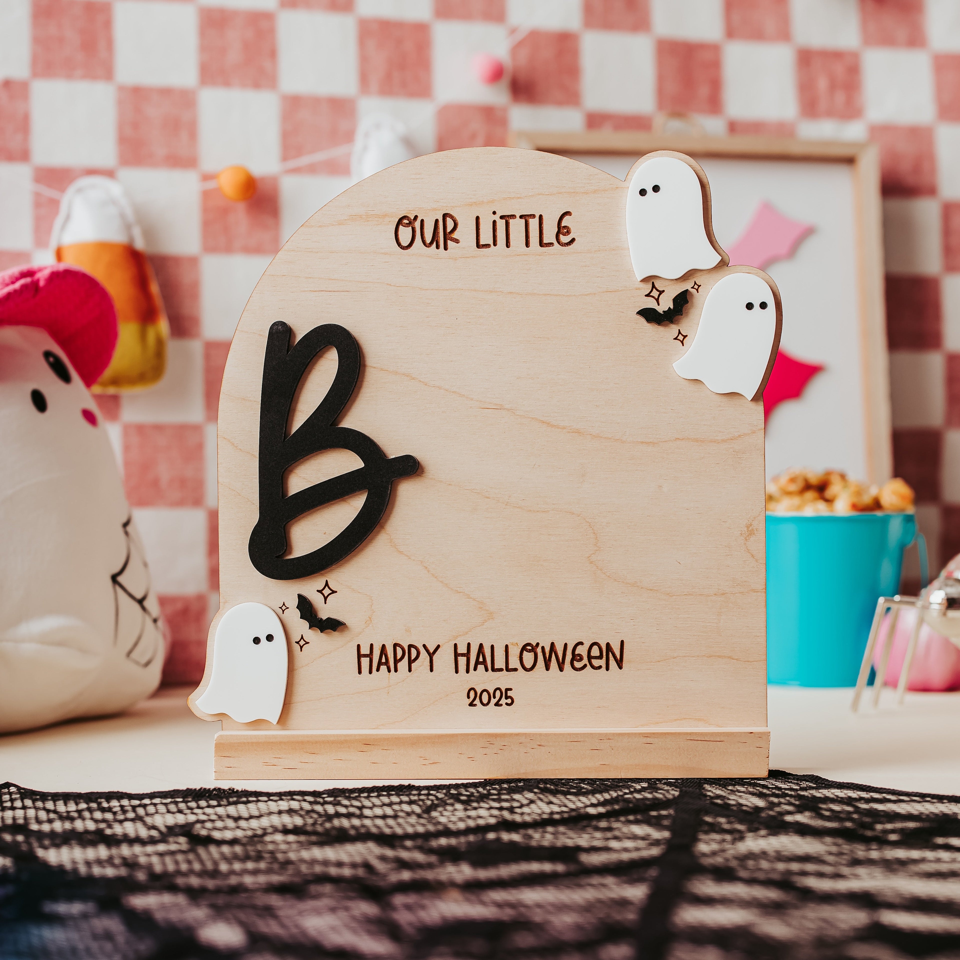A wooden Halloween tombstone decoration engraved with 'OUR LITTLE BOO HAPPY HALLOWEEN 2025', decorated with small white ghost cutouts, displayed on a decorated tabletop with candy corn and a checkered background.