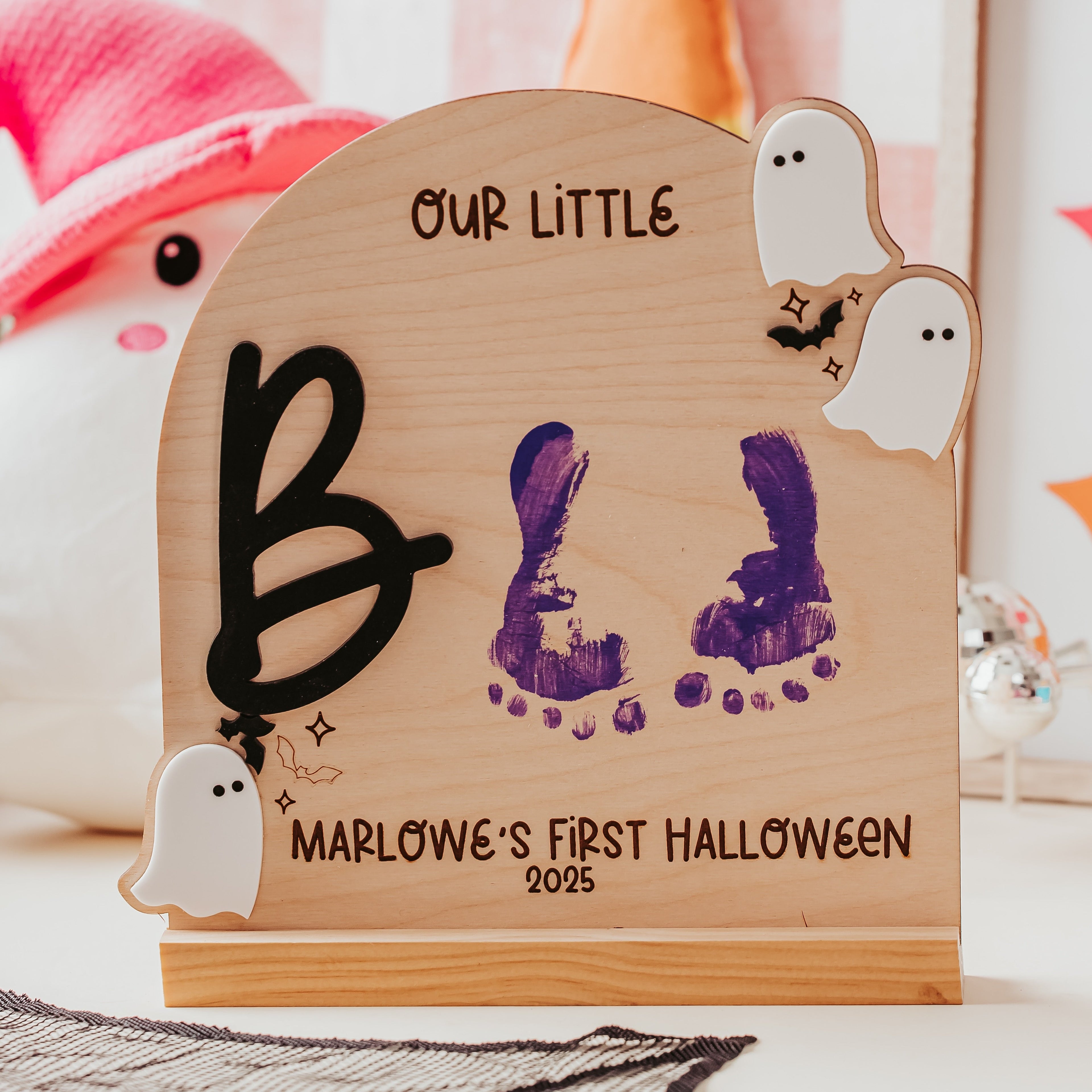 A personalized wooden Halloween plaque featuring a hand-painted purple bunny footprint, a black letter 'B', and ghost decorations, with the text 'OUR LITTLE BOO MARLOWE'S FIRST HALLOWEEN 2025'.