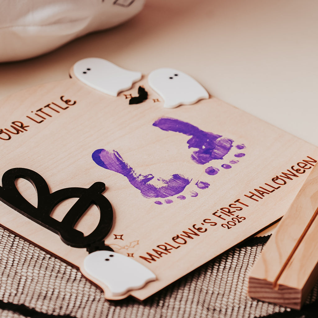 A wooden personalized Halloween keepsake board with a purple footprint print, ghost cutouts, and engraved text reading 'OUR LITTLE BOO MARLOWE'S FIRST HALLOWEEN 2026,' styled on a tabletop with a wooden block and black bat-wing decoration.