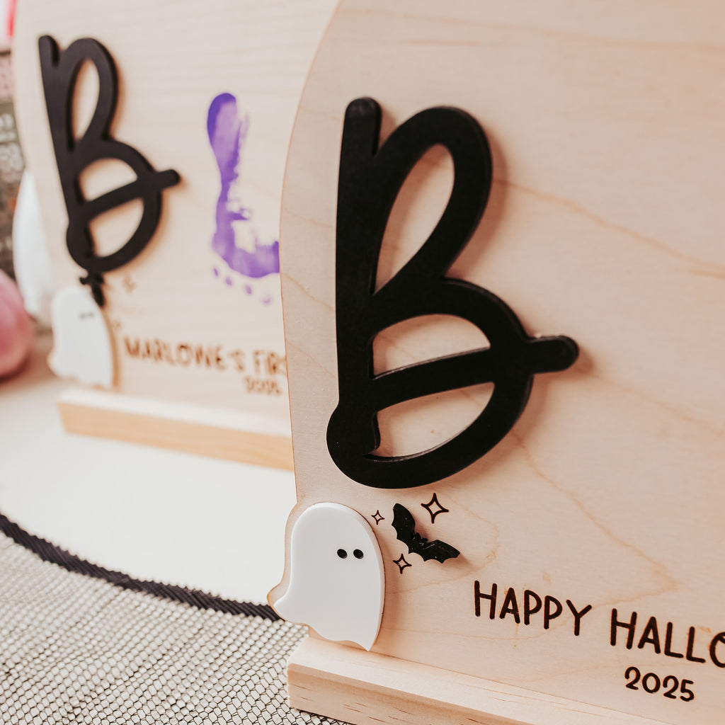 A close-up of a personalized wooden Halloween table sign with a large black letter 'B', a small white ghost, and 'HAPPY HALLOWEEN 2025' engraved on a light wood base. A matching sign is blurred in the background on a patterned surface.