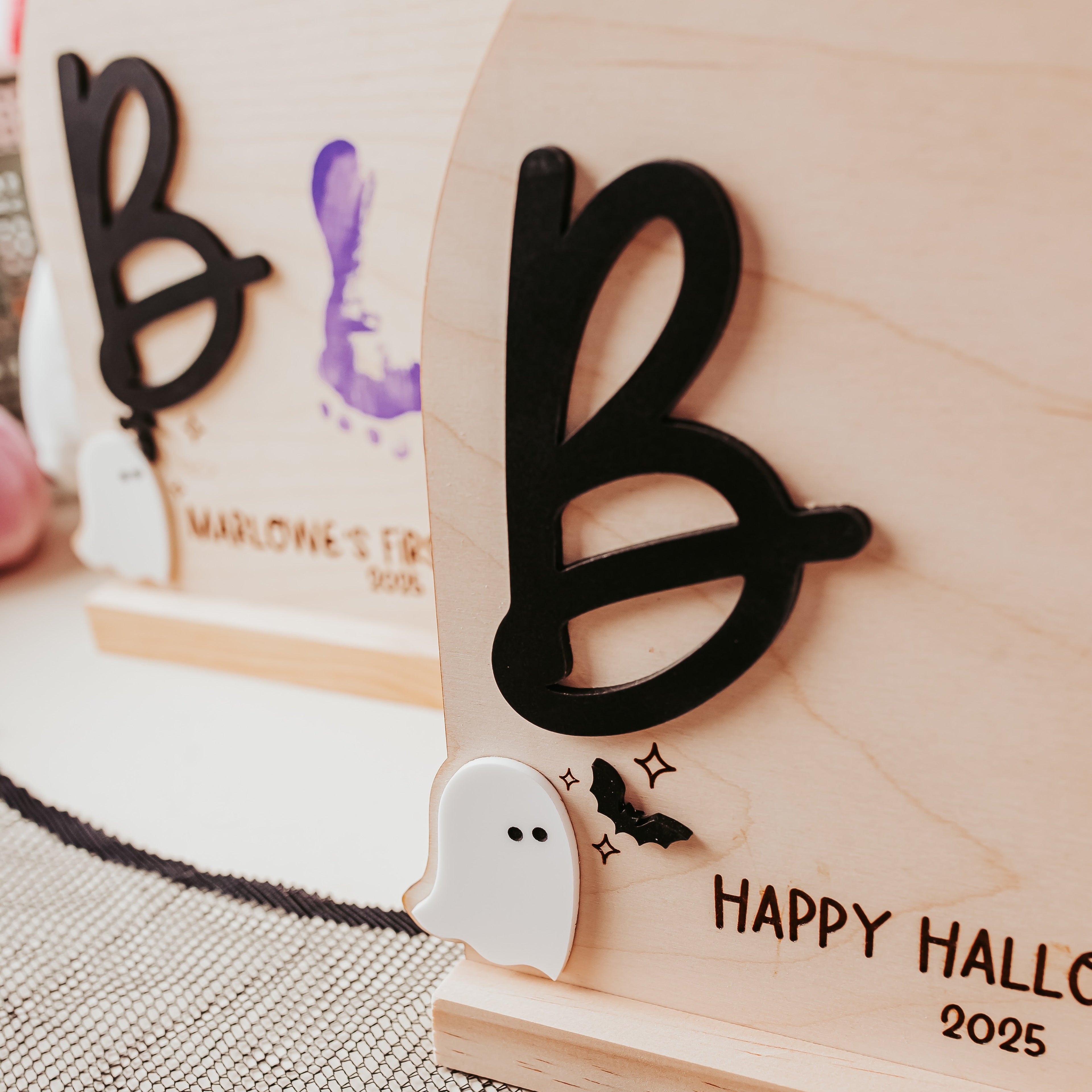 A close-up of a personalized wooden Halloween table sign with a large black letter 'B', a small white ghost, and 'HAPPY HALLOWEEN 2025' engraved on a light wood base. A matching sign is blurred in the background on a patterned surface.