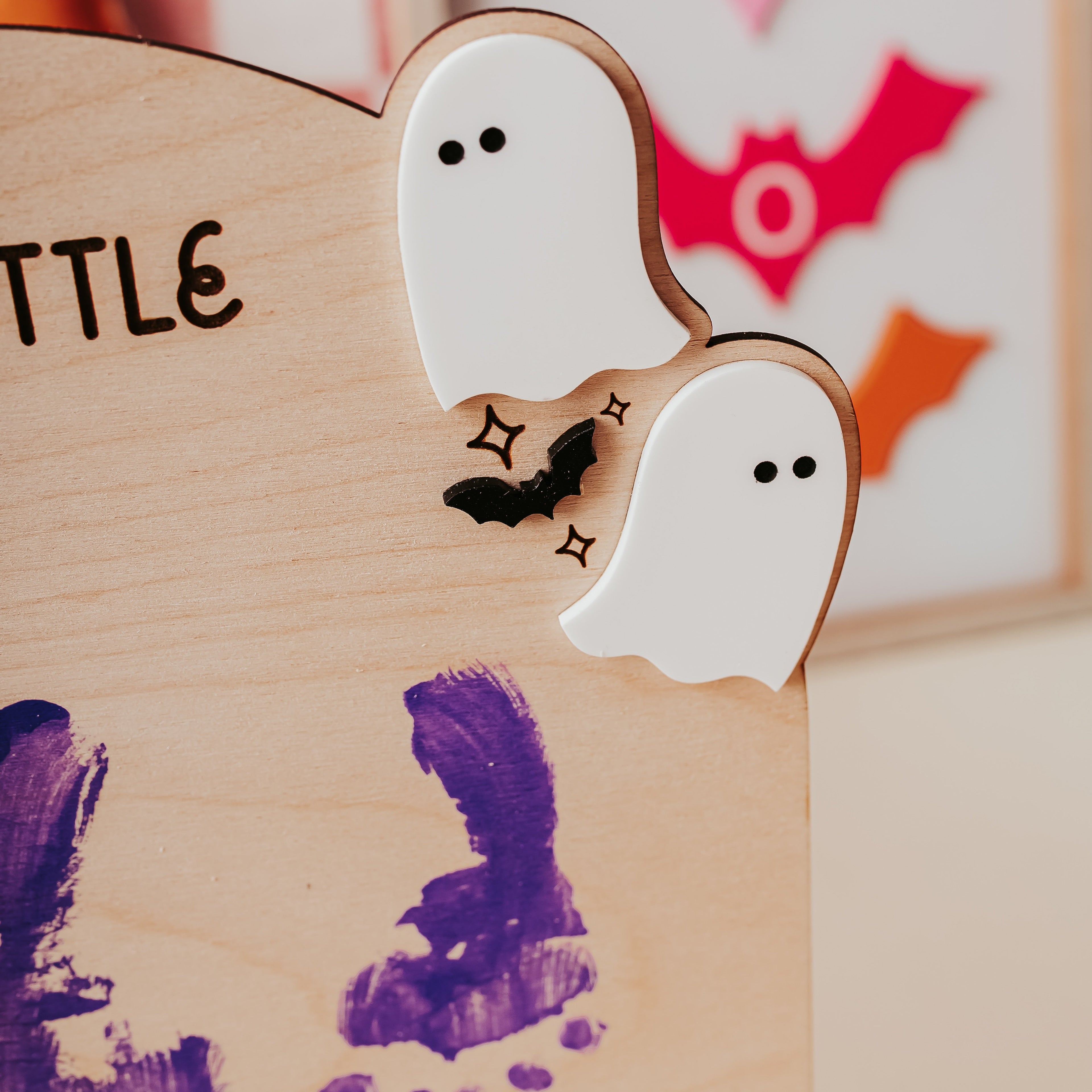 Close-up of a wooden children's display board with two white ghost cutouts, a small black bat, and purple handprint decorations, with a blurred pink-and-white framed bat artwork in the background.