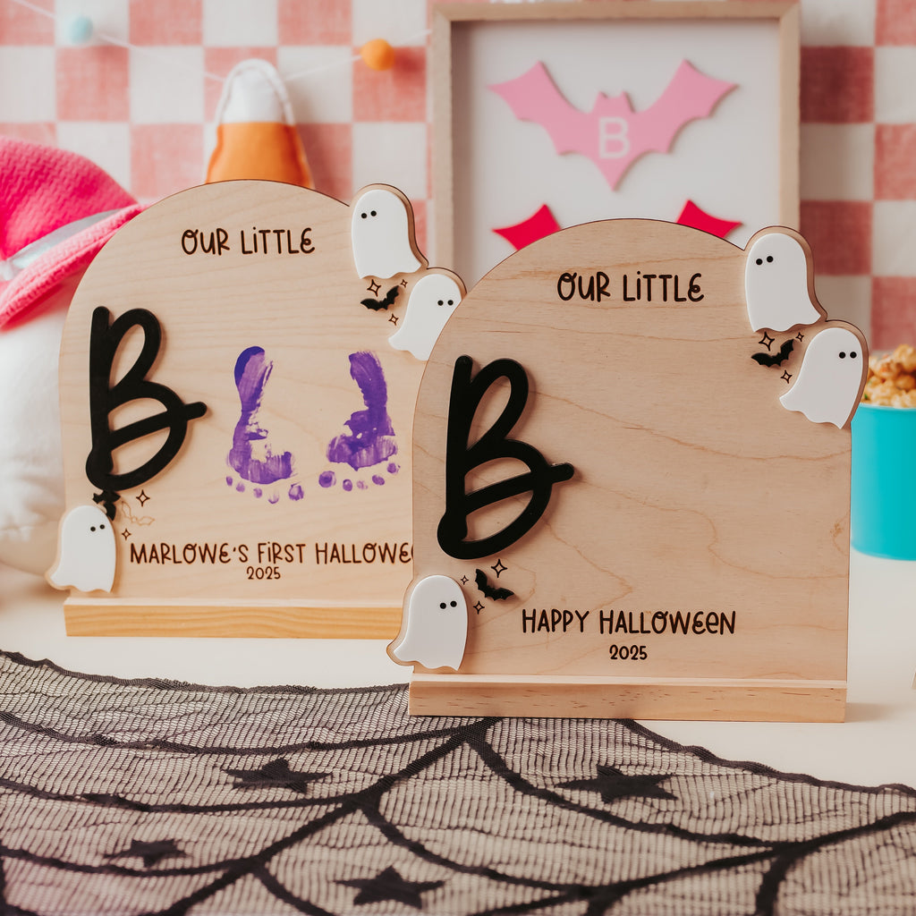 Two personalized wooden Halloween plaques displayed on a decorated tabletop. The plaques are light natural wood with black lettering that reads 'OUR LITTLE' and a large black letter 'B', with one plaque showing purple baby footprints and the other reading 'HAPPY HALLOWEEN 2025'.