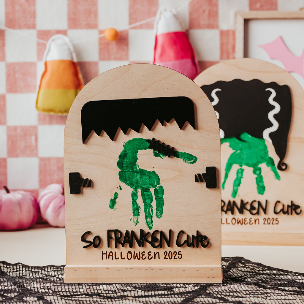 Two wooden Halloween arch decorations displayed on a black lace surface, with a pink-and-white checkered backdrop and candy corn props in the background. The front tombstone reads 'So FRANKEN Cute HALLOWEEN 2025' and features a green handprint with black stitch detail.