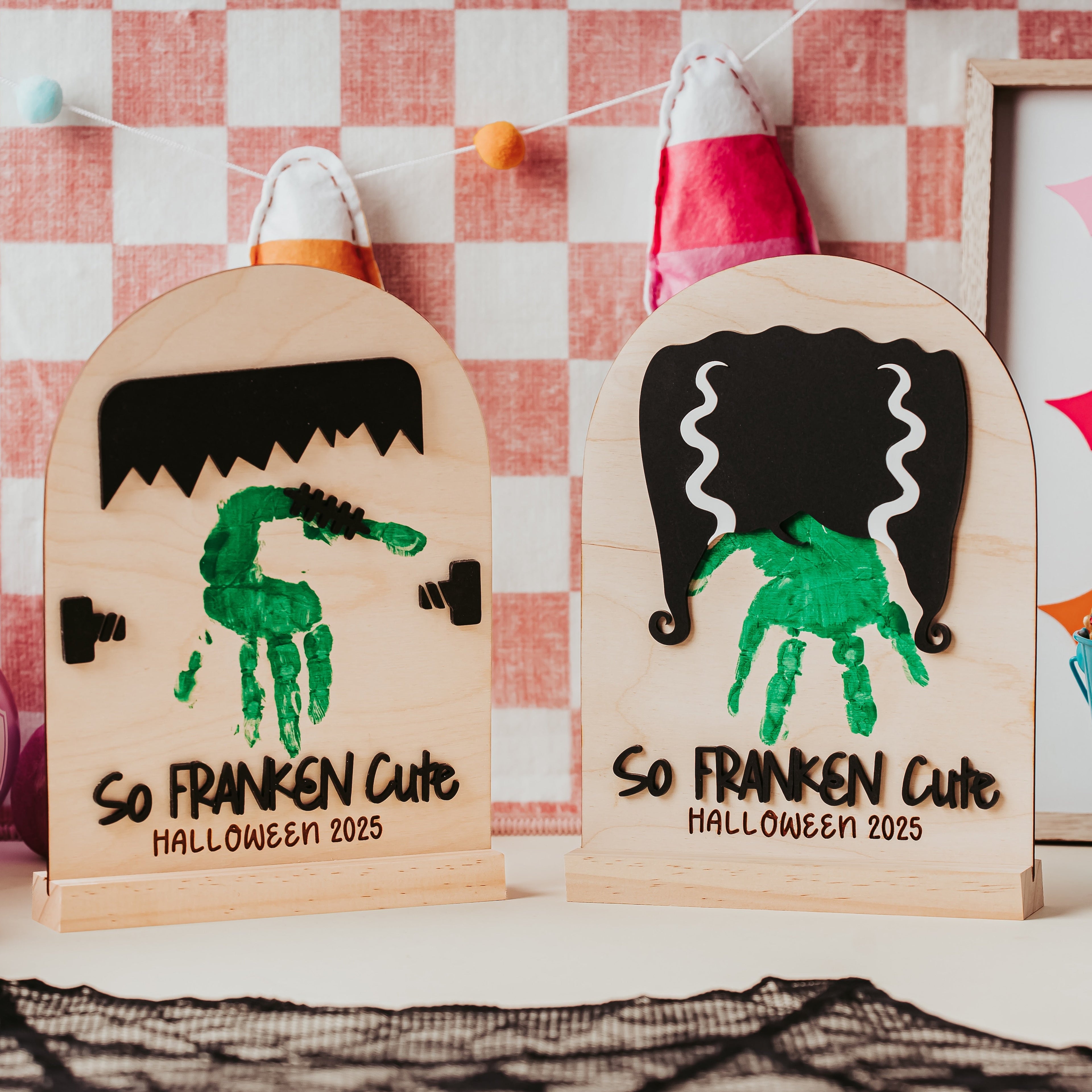 Two personalized wooden Halloween arch signs with green handprint graphics and the text 'So FRANKEN Cute HALLOWEEN 2025' displayed on a tabletop with festive decorations in the background.