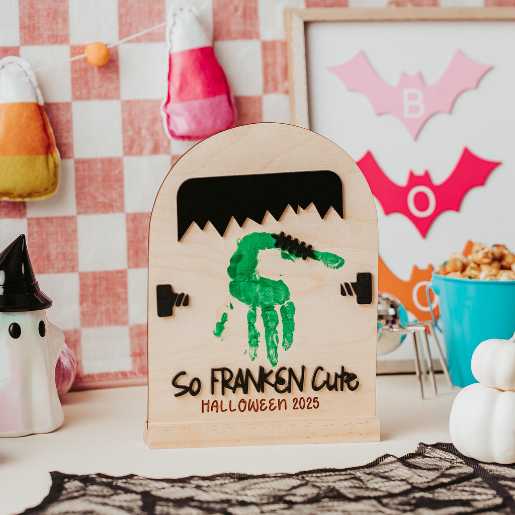 A personalized wooden Halloween handprint sign with a green handprint and the text 'SO FRANKEN CUTE HALLOWEEN 2025', styled on a tabletop with candy corn, a ghost figurine, and a checkered wall background.