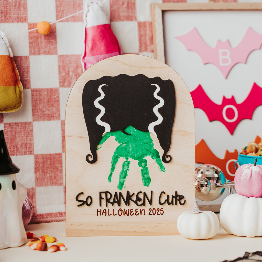 A personalized wooden Halloween sign featuring a green handprint under black hair cutouts, with the text 'So FRANKEN Cute HALLOWEEN 2025'. The sign is styled on a tabletop with candy corn, small pumpkins, a framed bat cutout, and a red-and-white checkered background.