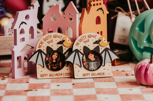 Two Halloween-themed wooden arch-shaped photo magnets with bat wings and circular cutouts, displayed on a checkered tablecloth with other seasonal decor in the background.