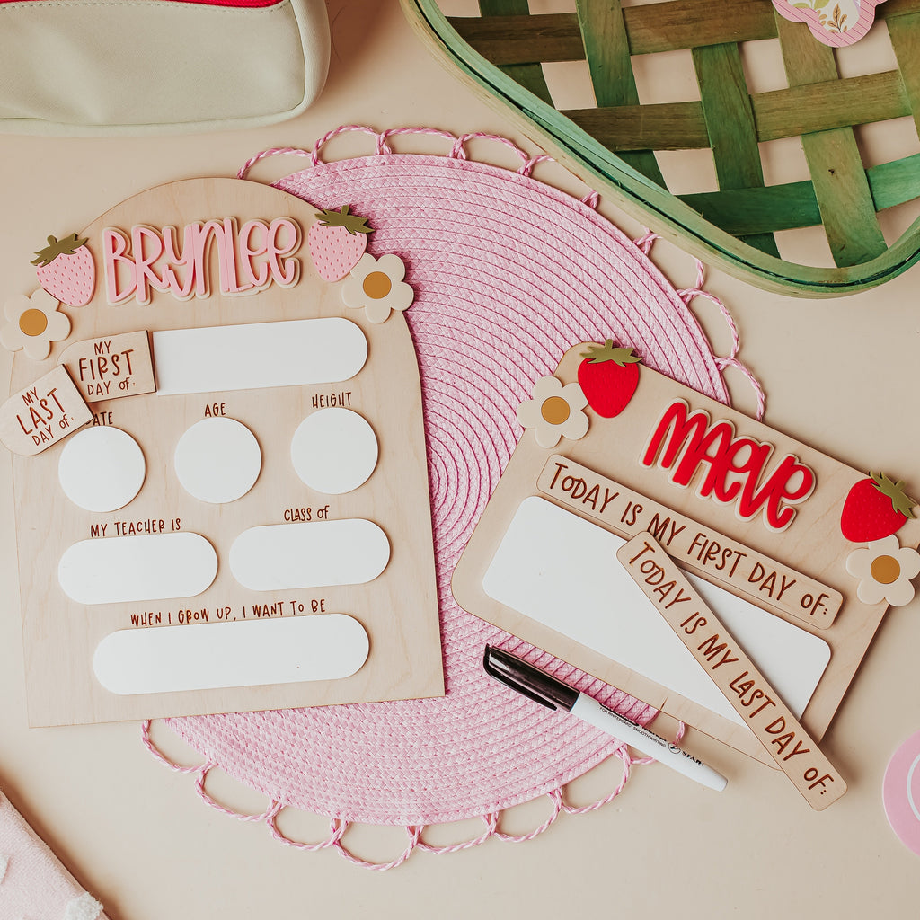 Two wooden first-day-of-school cry erase signs with pink and red strawberries, styled on a pink woven placemat with pastel stationery props on a beige background.