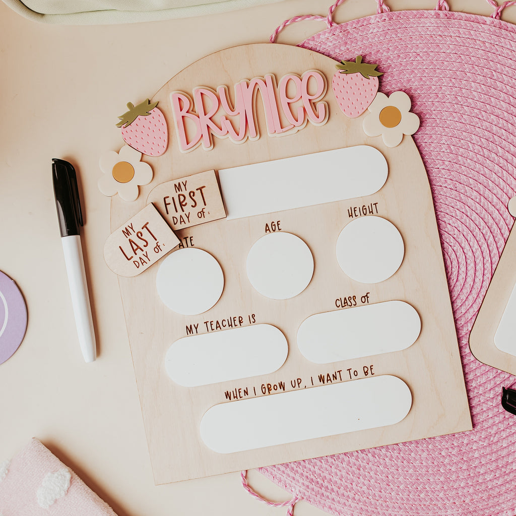 A personalized wooden back to school dry erase styled in a flat-lay arrangement on a light pink background, surrounded by a cream zippered pouch, a pink woven mat, a black marker, and a pink cloud notebook.
