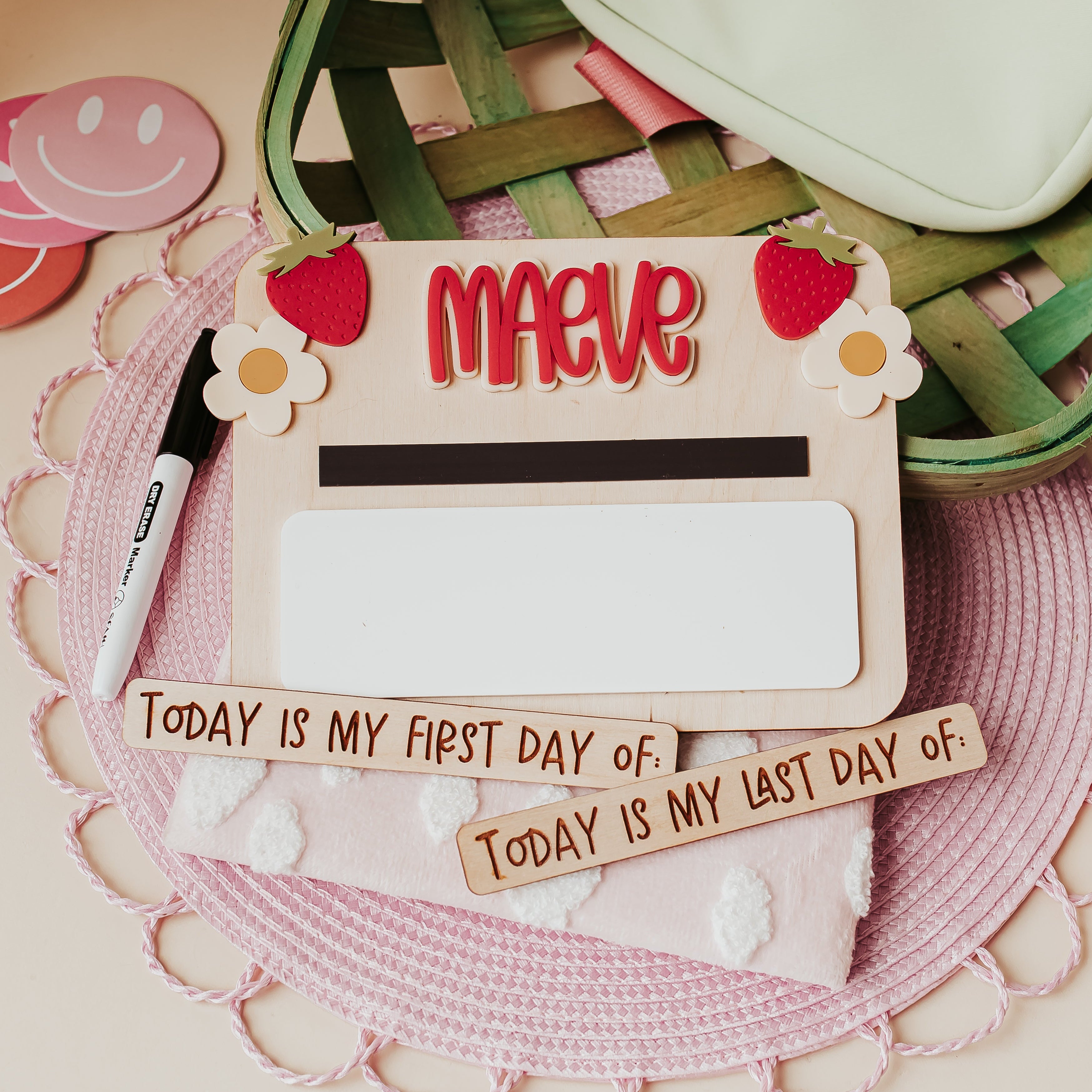 A personalized wooden first-day-of-school sign with the name 'MAEVE' in red letters, decorated with strawberries and flowers. The board is styled on a pink lace mat with a marker, a pink pouch, and wooden text plaques that read 'TODAY IS MY FIRST DAY OF:' and 'TODAY IS MY LAST DAY OF:'.