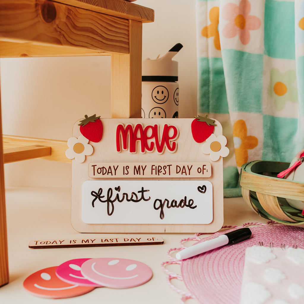 A personalized first-day-of-school sign reading 'Maeve - today is my first day of first grade' styled on a desk with pink smiley face coasters, a pink marker, a patterned curtain, and a wooden shelf in the background.
