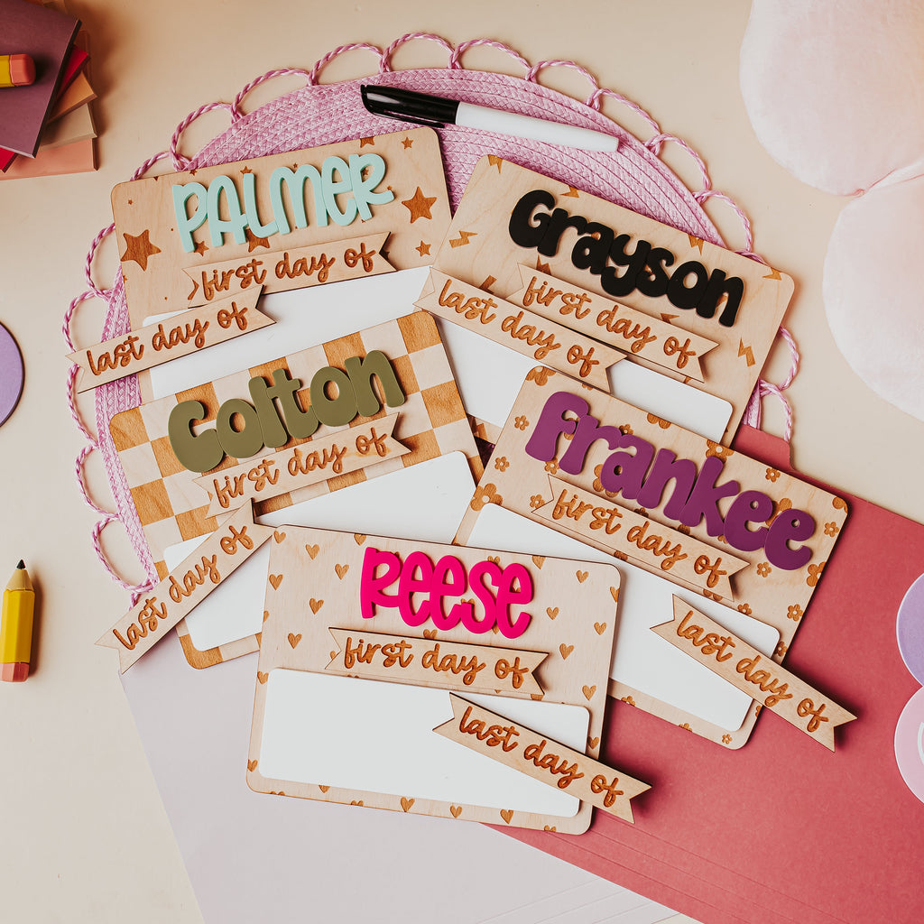 A styled flat-lay of personalized first-day-of-school dry erase signs on a light beige surface with a pink circular doily, a dry erase marker, a pink folder , and pastel smiley face post-its.