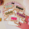 A styled flat-lay of personalized first-day-of-school dry erase signs on a light beige surface with a pink circular doily, a dry erase marker, a pink folder , and pastel smiley face post-its.