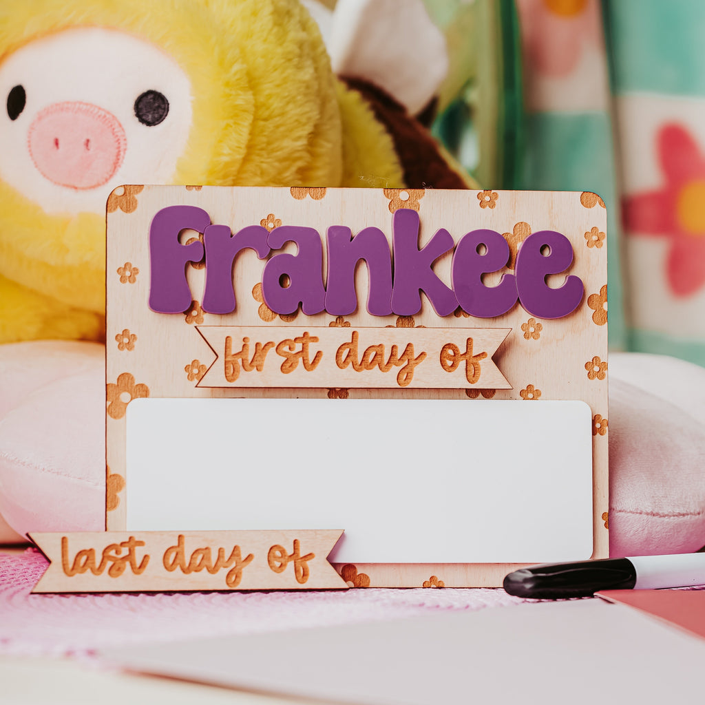 A decorative wooden back to school dry erase sign with engraved flowers and  the name 'frankee' in large purple letters and banners reading 'first day of' and 'last day of', styled with a plush toy and stationery in the background.