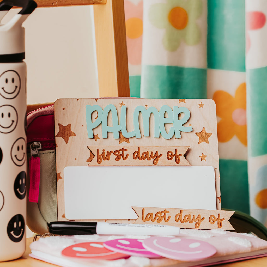 A personalized wooden first-day-of-school sign with the name 'PALMER' in blue and 'first day of' in brown script, styled on a wooden desk with a smiley-face water bottle, notebook, and flower-patterned curtain in the background.