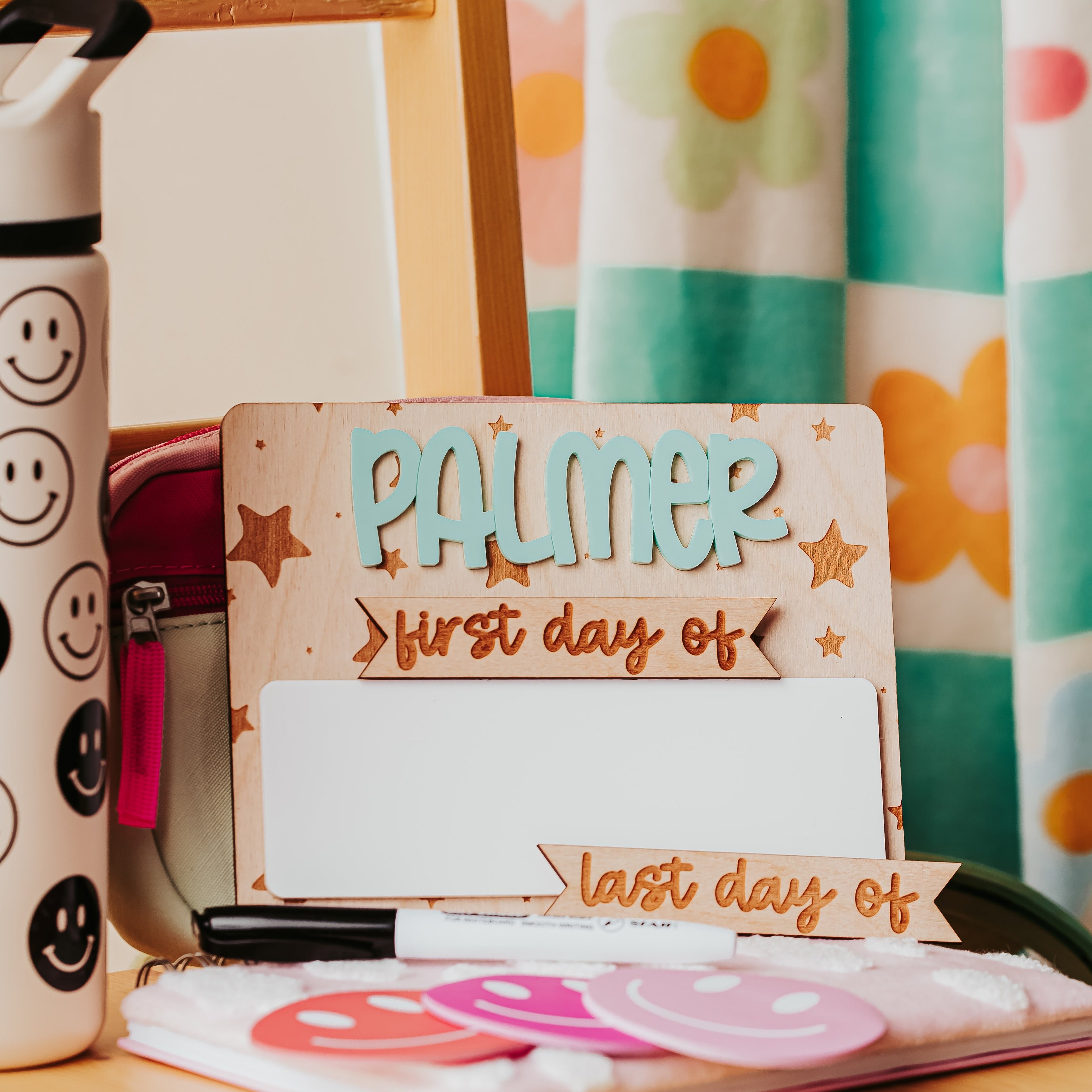 A personalized wooden first-day-of-school sign with the name 'PALMER' in blue and 'first day of' in brown script, styled on a wooden desk with a smiley-face water bottle, notebook, and flower-patterned curtain in the background.