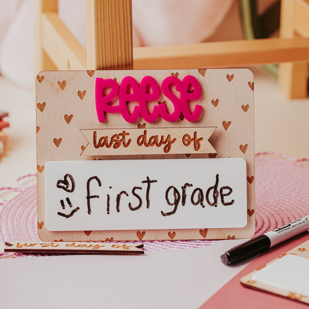 A personalized first-day-of-school sign with the name 'Reese' in bright pink letters and 'last day of first grade' on a wooden stand, styled on a desk with a marker and pink paper.