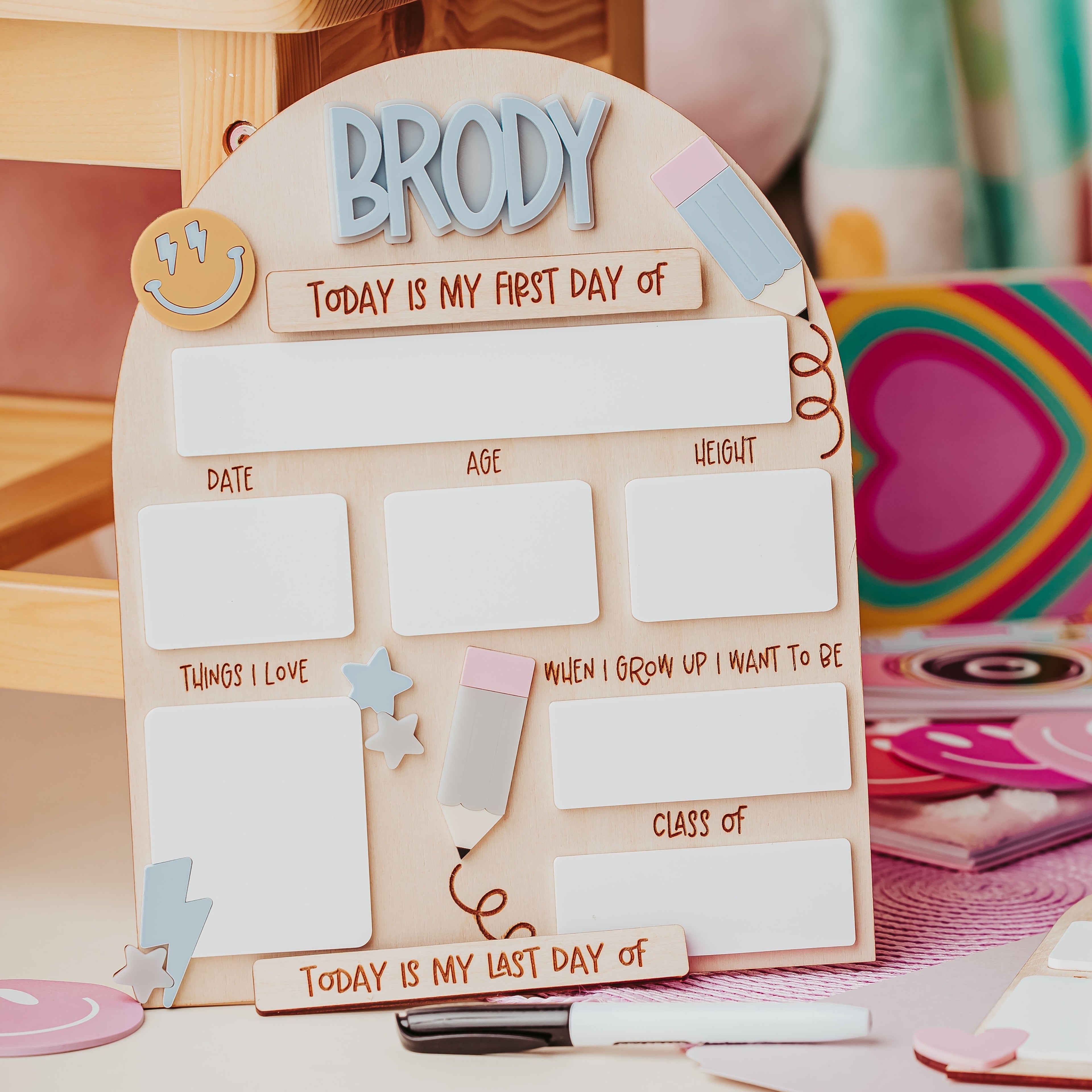 A personalized wooden first-day-of-school dry erase board board with the name 'Brody' at the top, set on a desk with school supplies in a cozy, pastel-colored room.