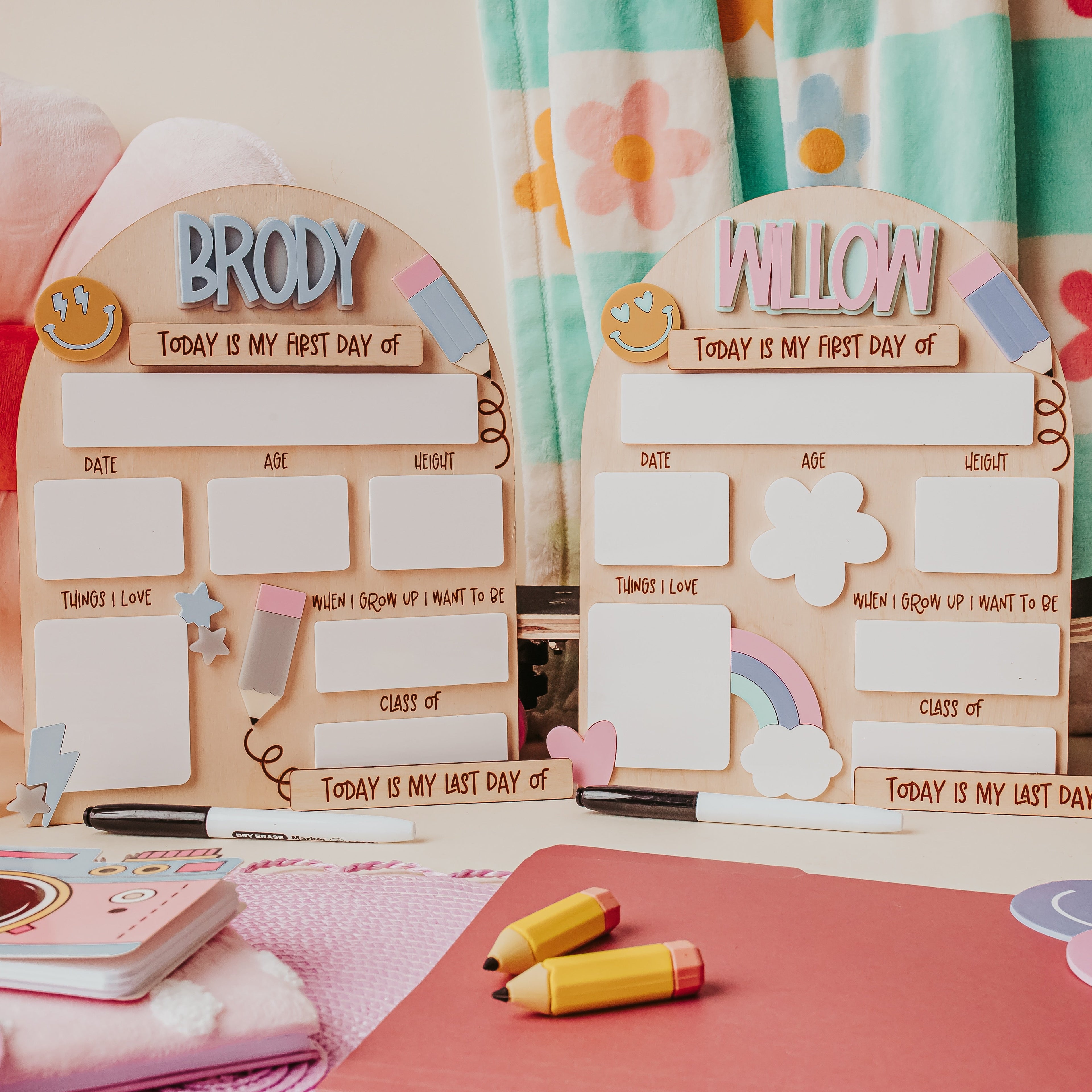 Two personalized wooden first-day-of-school back to school dry erase signs  labeled BRODY and WILLOW, styled on a desk with a pink notebook, yellow markers, and colorful accessories in a room setting.