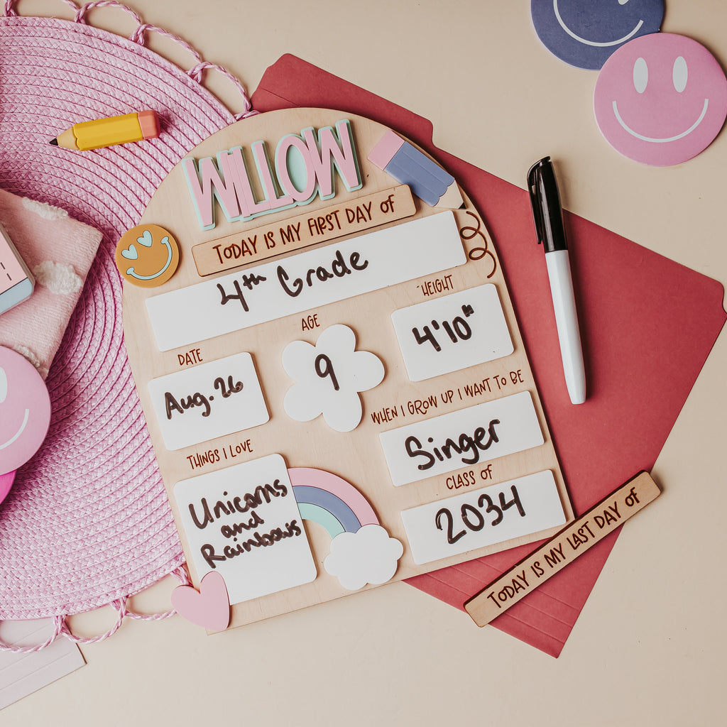 A styled flat-lay of a personalized wooden first-day-of-school sign with cute pastel stickers, a pink camera illustration, smiley face cutouts, a red envelope, and a black marker on a beige background.
