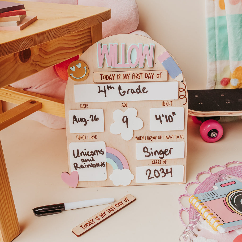 A decorative wooden dry erase back to school board styled on a desk with a chalk, notebook, and other school-themed props. The plaque is personalized with the name 'WILLOW', 'TODAY IS MY FIRST DAY OF 4th Grade', and various date and milestone notes.