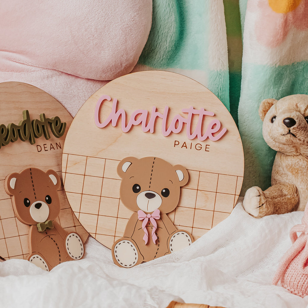 Personalized wooden name signs featuring teddy bear illustrations, styled in a soft baby-themed setting with a pink plush toy, mint and pink floral blanket, and a wicker basket.