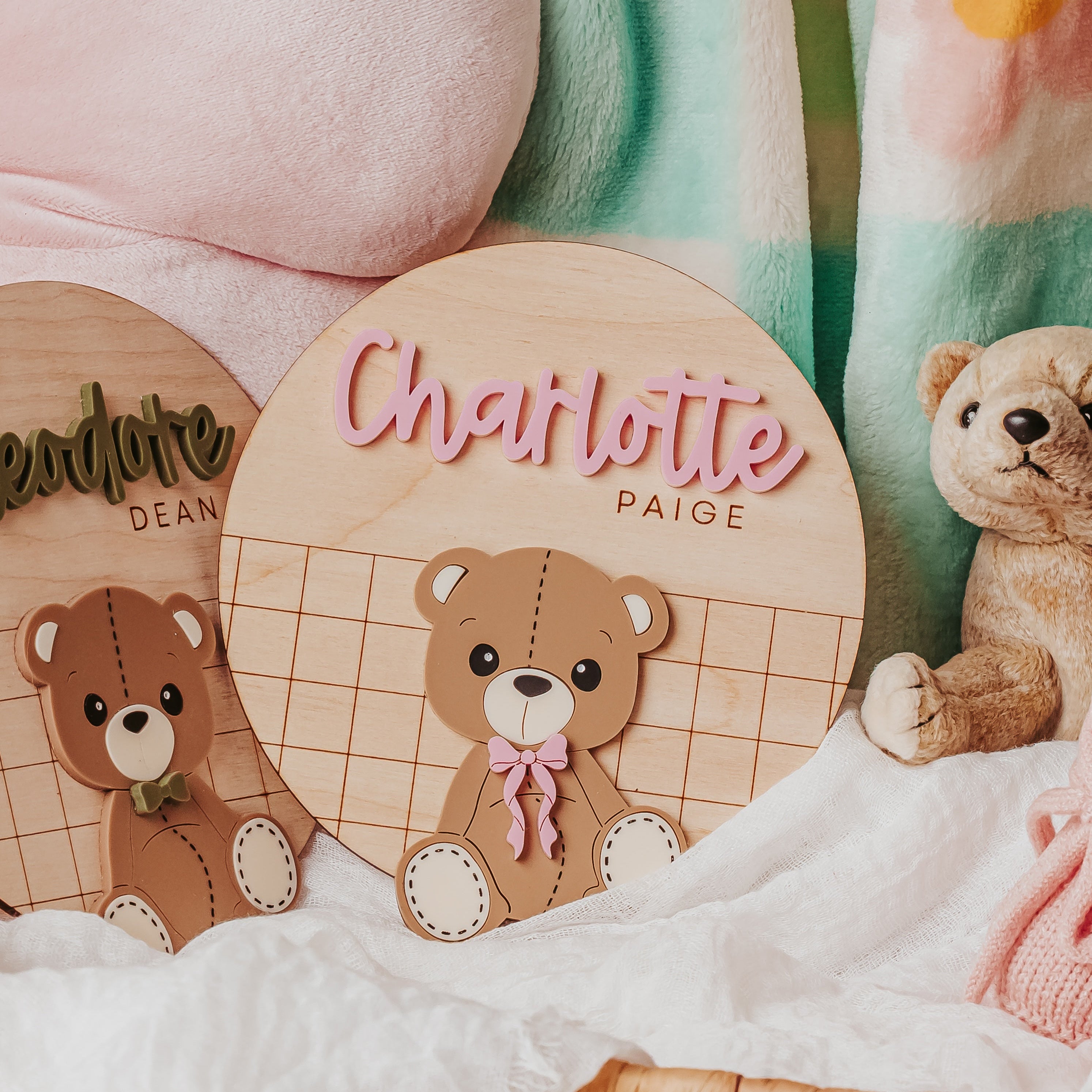 Personalized wooden name signs featuring teddy bear illustrations, styled in a soft baby-themed setting with a pink plush toy, mint and pink floral blanket, and a wicker basket.