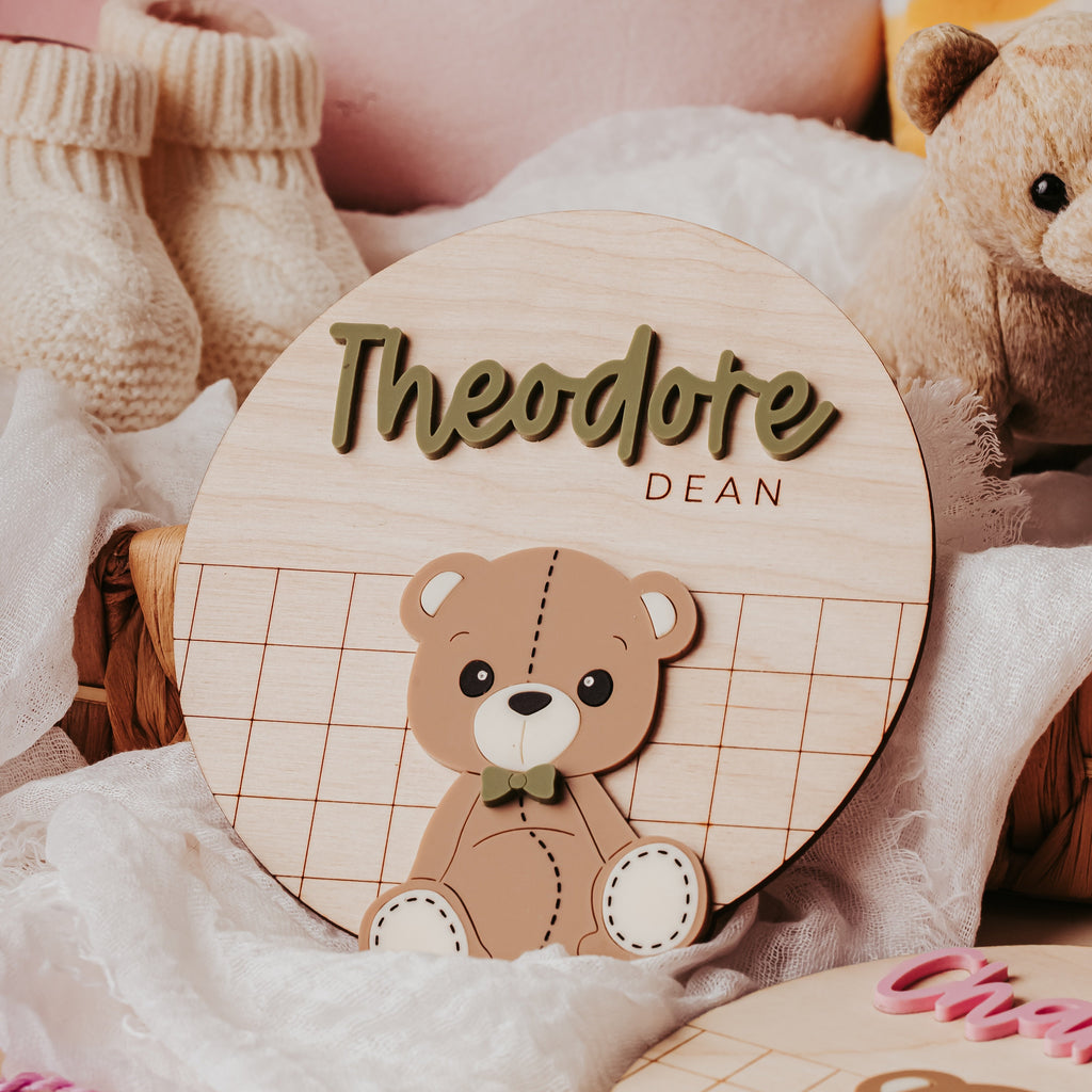 A personalized wooden baby name sign keepsake featuring a cute teddy bear 3D illustration and the name 'Theodote' with 'DEAN' underneath, styled in a cozy nursery setting with plush toys, knit booties, and soft blankets.