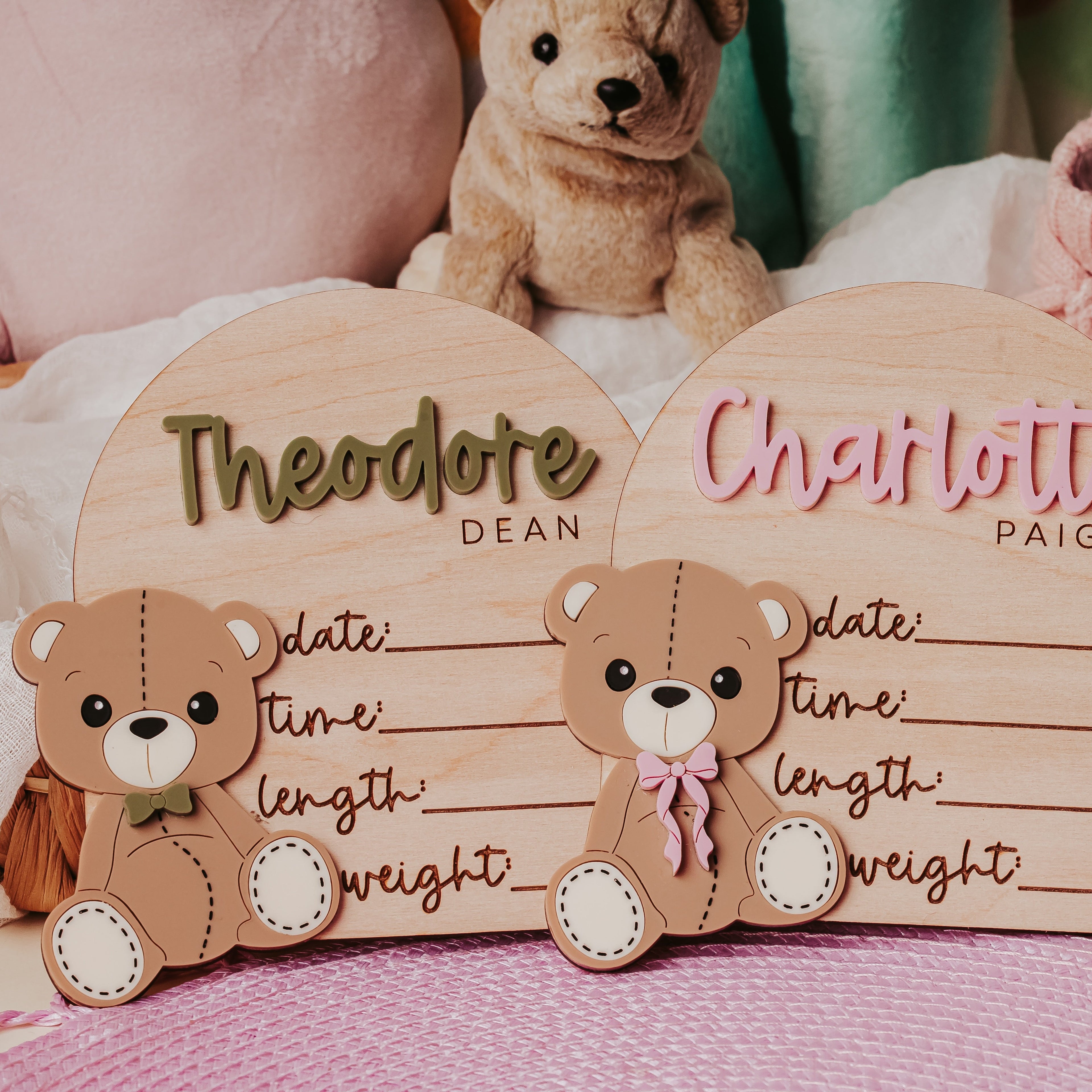 Two personalized wooden baby name signs shaped like arched cards with 3D teddy bears, displayed in a styled nursery setting with a stuffed bear, pink booties, and pillows.