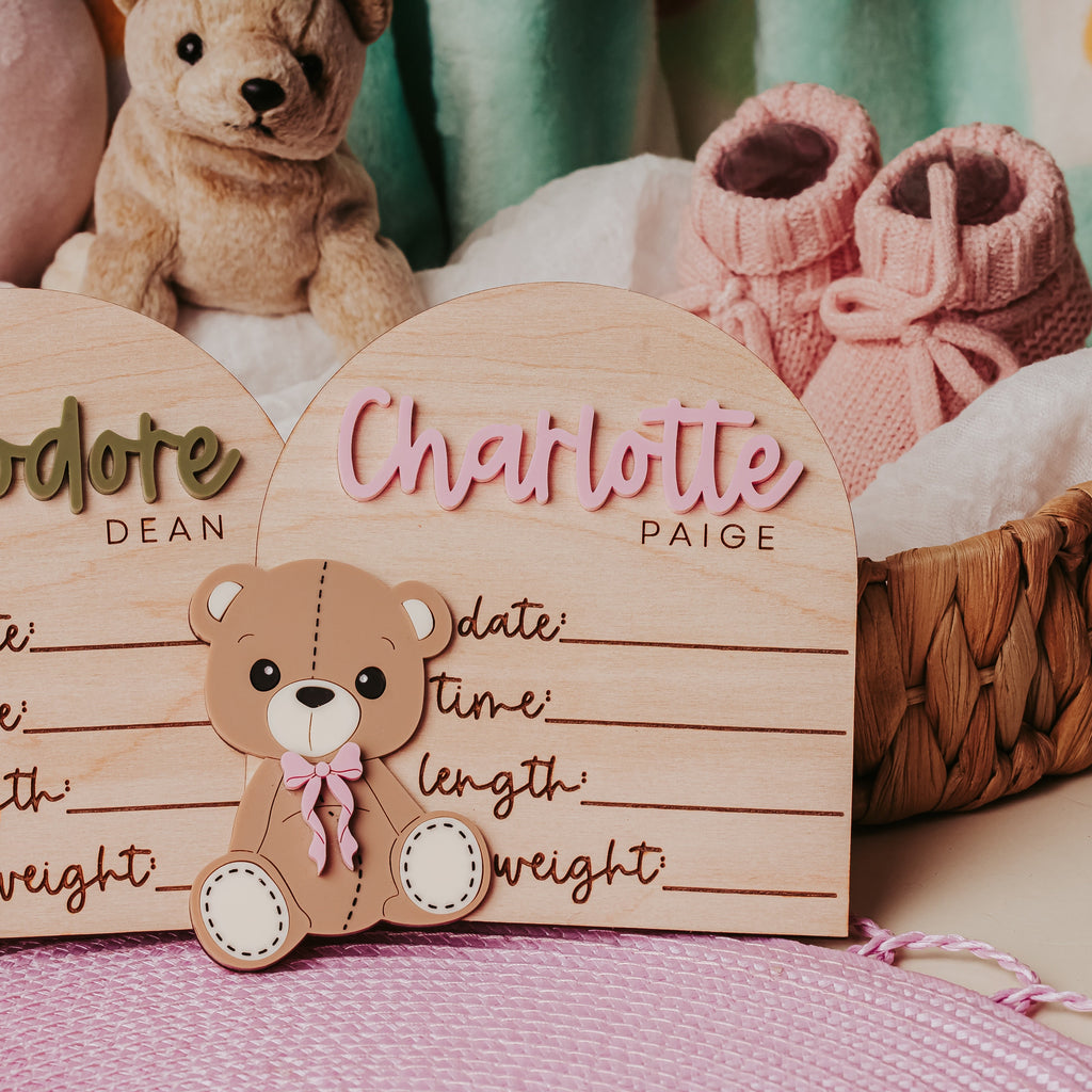 Two personalized wooden baby name signs shaped like arched cards with teddy bear cutouts, displayed in a styled nursery setting with a stuffed bear, pink booties, and pillows.