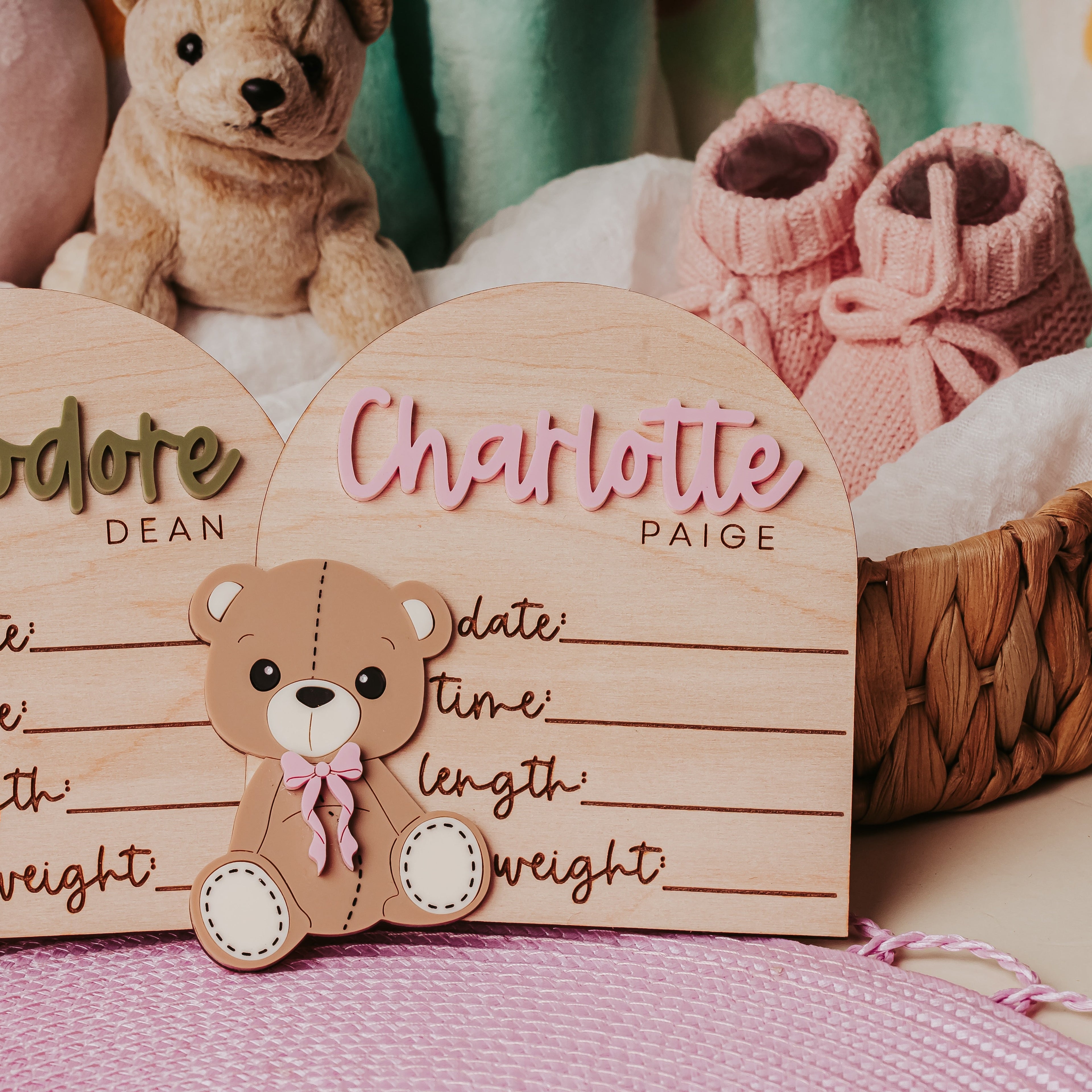Two personalized wooden baby name signs shaped like arched cards with teddy bear cutouts, displayed in a styled nursery setting with a stuffed bear, pink booties, and pillows.