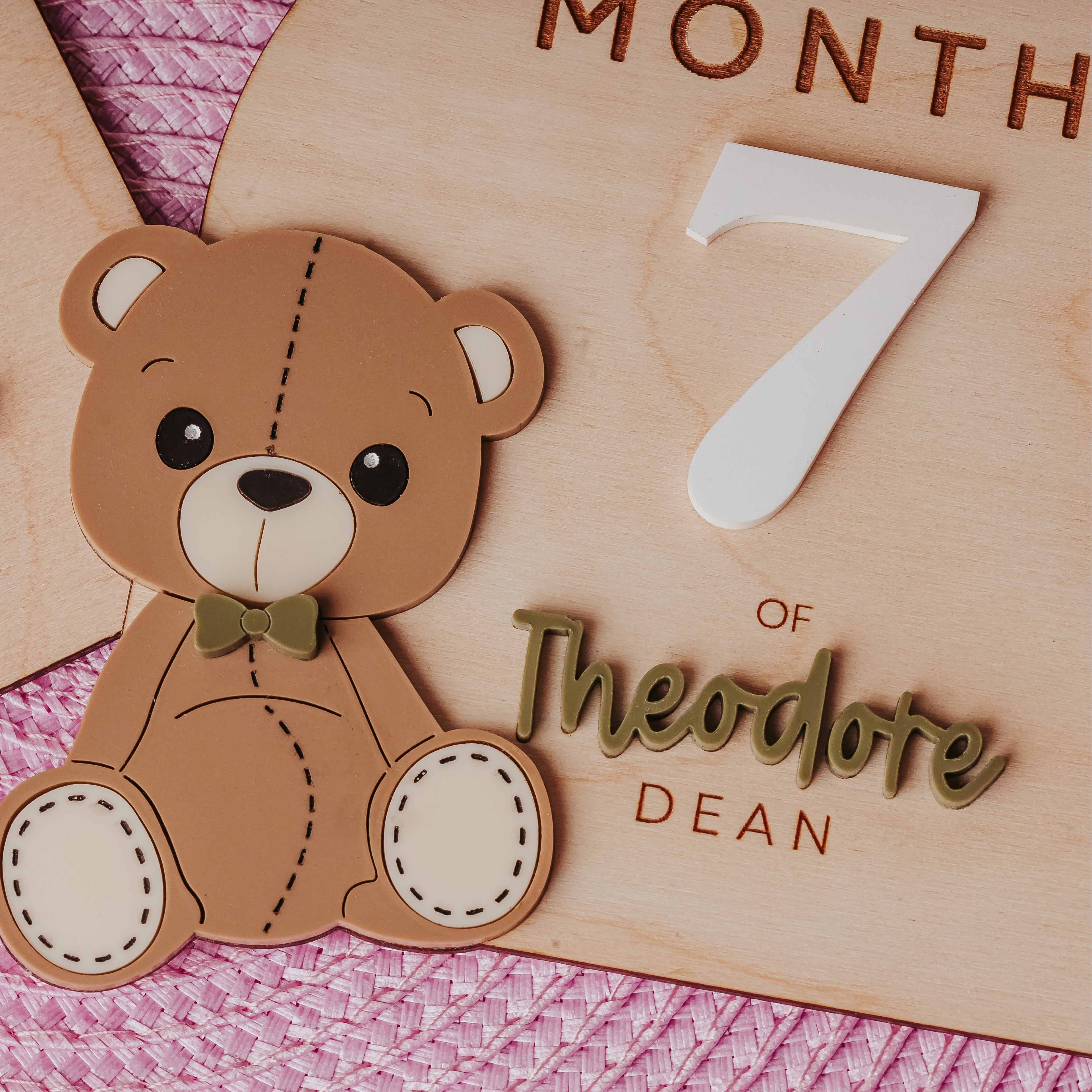Personalized wooden baby milestone plaques featuring a cute brown teddy bear with a bow tie, displayed on a pink textured blanket. 