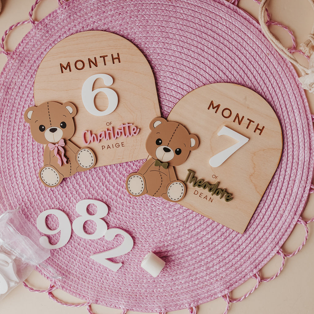 A baby milestone plaque set featuring two wooden arch plaques labeled 'MONTH 6' and 'MONTH 7' with custom names, two small teddy bear cutouts, and white numbers '982' arranged on a pink woven placemat.