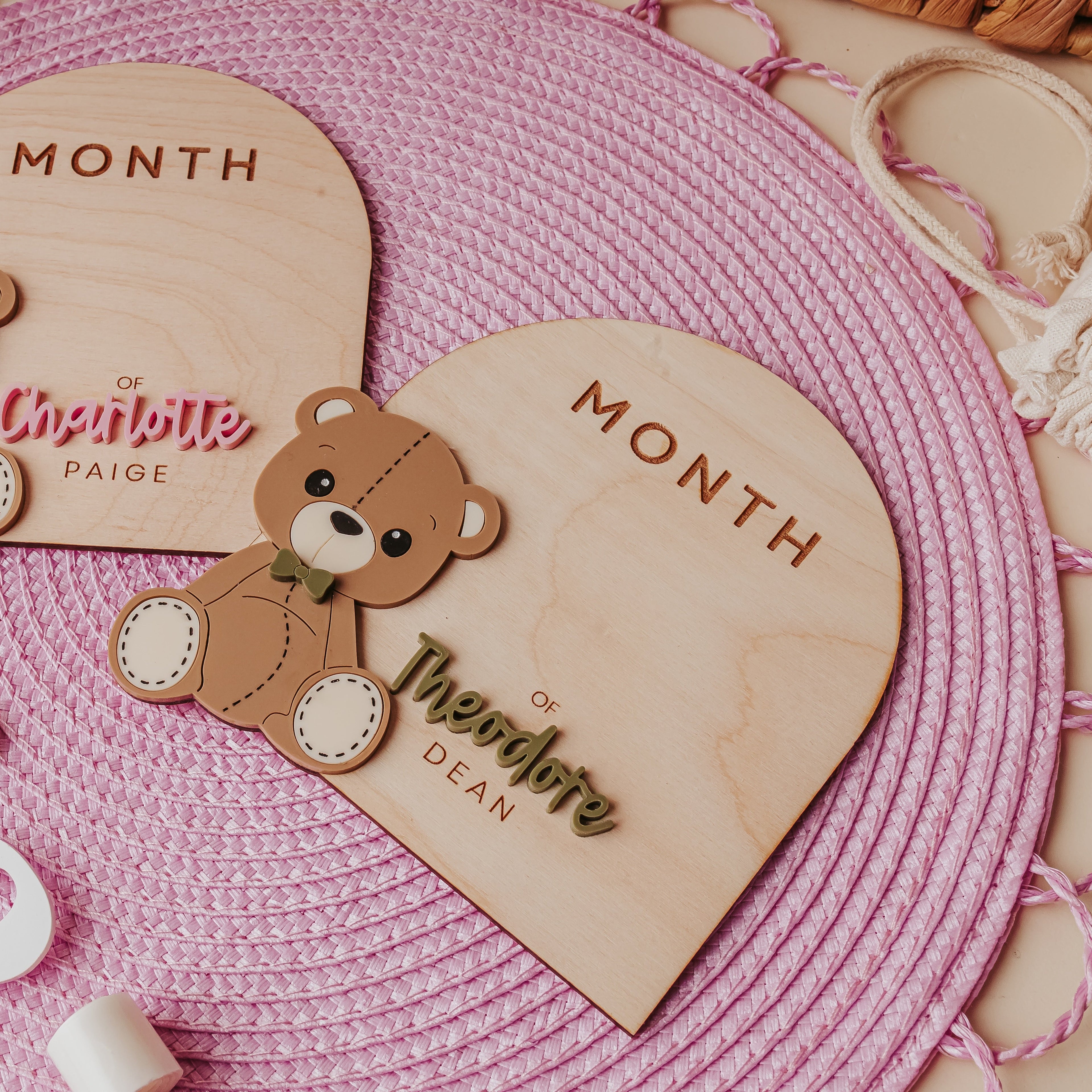 Two personalized wooden baby milestone plaques shaped like teddy bears, displayed on a pink woven placemat with white decorative numbers.