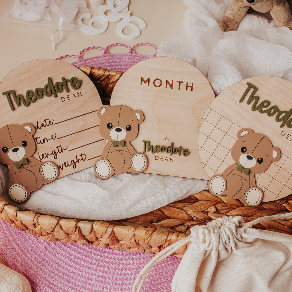 Three wooden baby keepsake tags with 3D teddy bear illustrations are displayed in a pink woven basket with white fabric and baby booties.