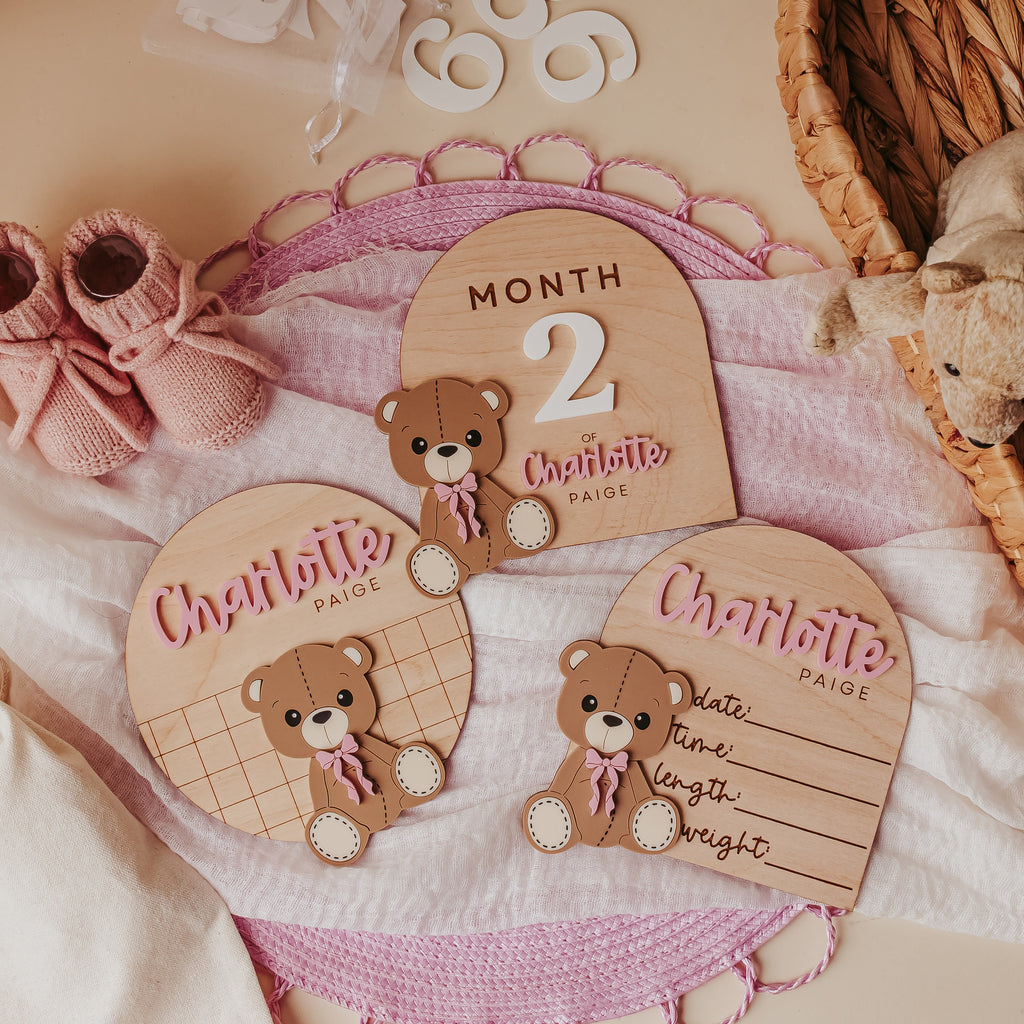 A styled flat-lay of personalized wooden baby milestone plaques, baby name announcement and baby birth stat on a soft pink blanket, with pink knit booties, white numerals, and a woven basket in the background.