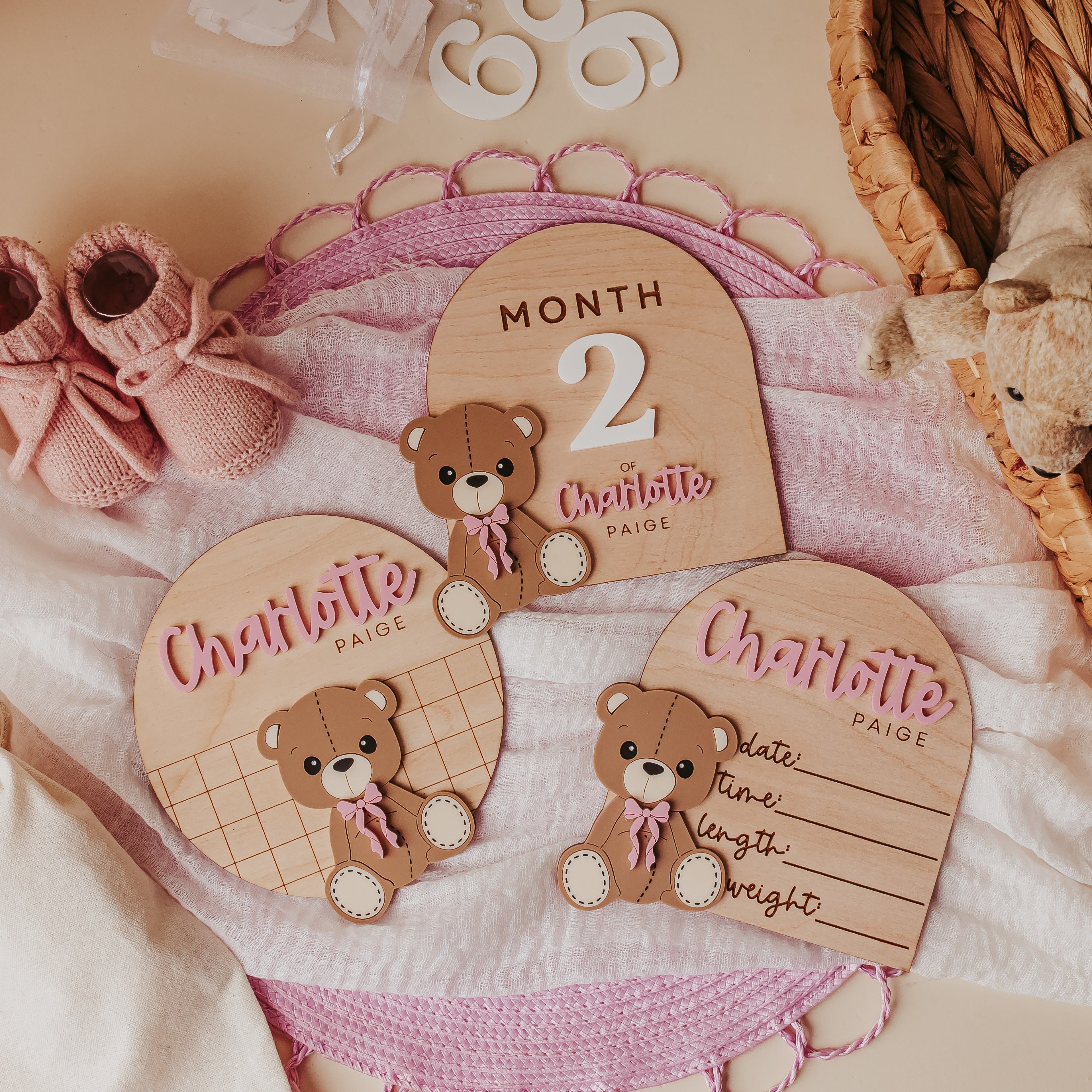 A styled flat-lay of personalized wooden baby milestone plaques, baby name announcement and baby birth stat on a soft pink blanket, with pink knit booties, white numerals, and a woven basket in the background.