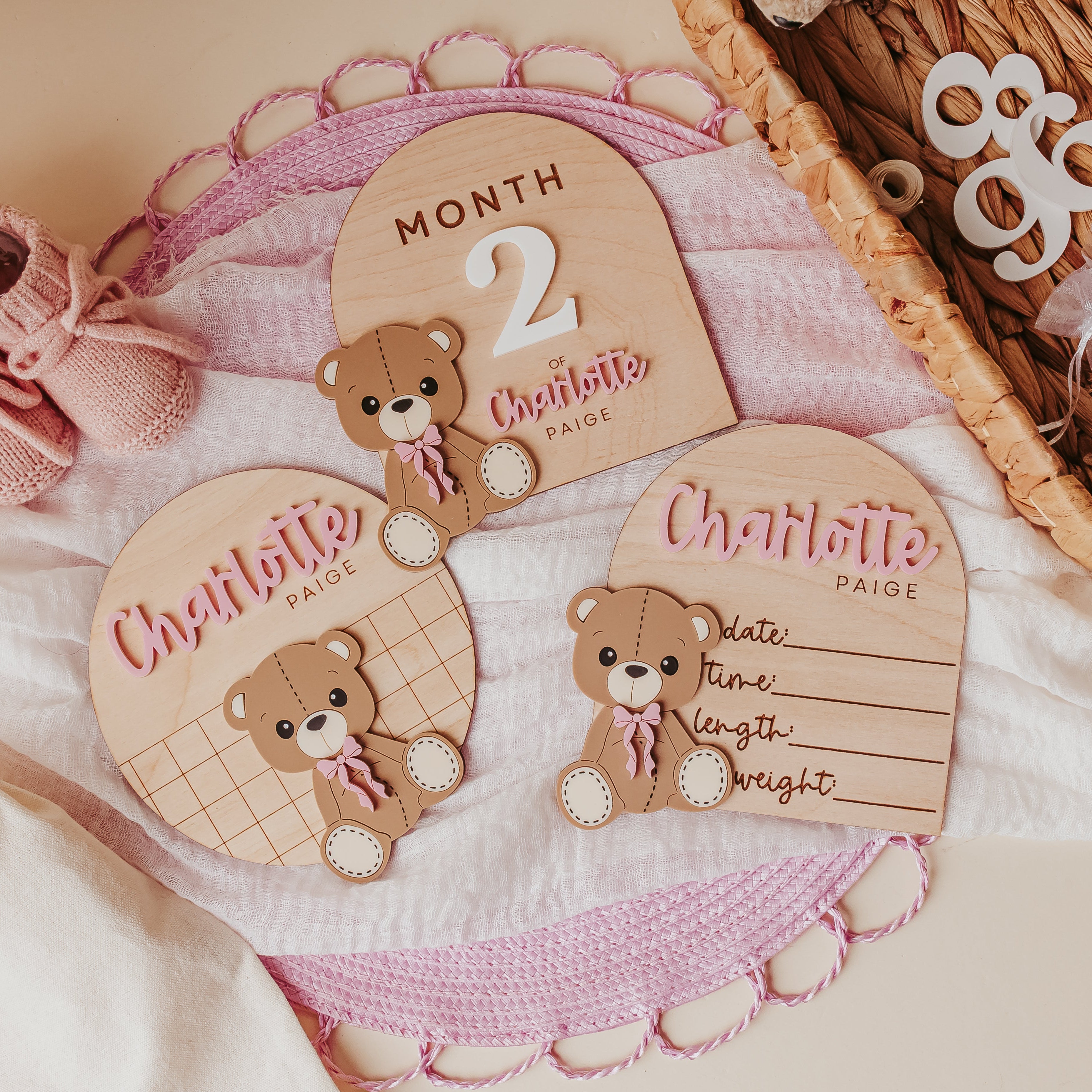 A styled flat-lay of personalized baby milestone cards and matching baby booties on soft white fabric. The set includes three wooden arch-shaped and oval plaques with teddy bear illustrations and pink script text reading names and months, alongside a pair of pink knit booties and a woven basket with white baby clothes.