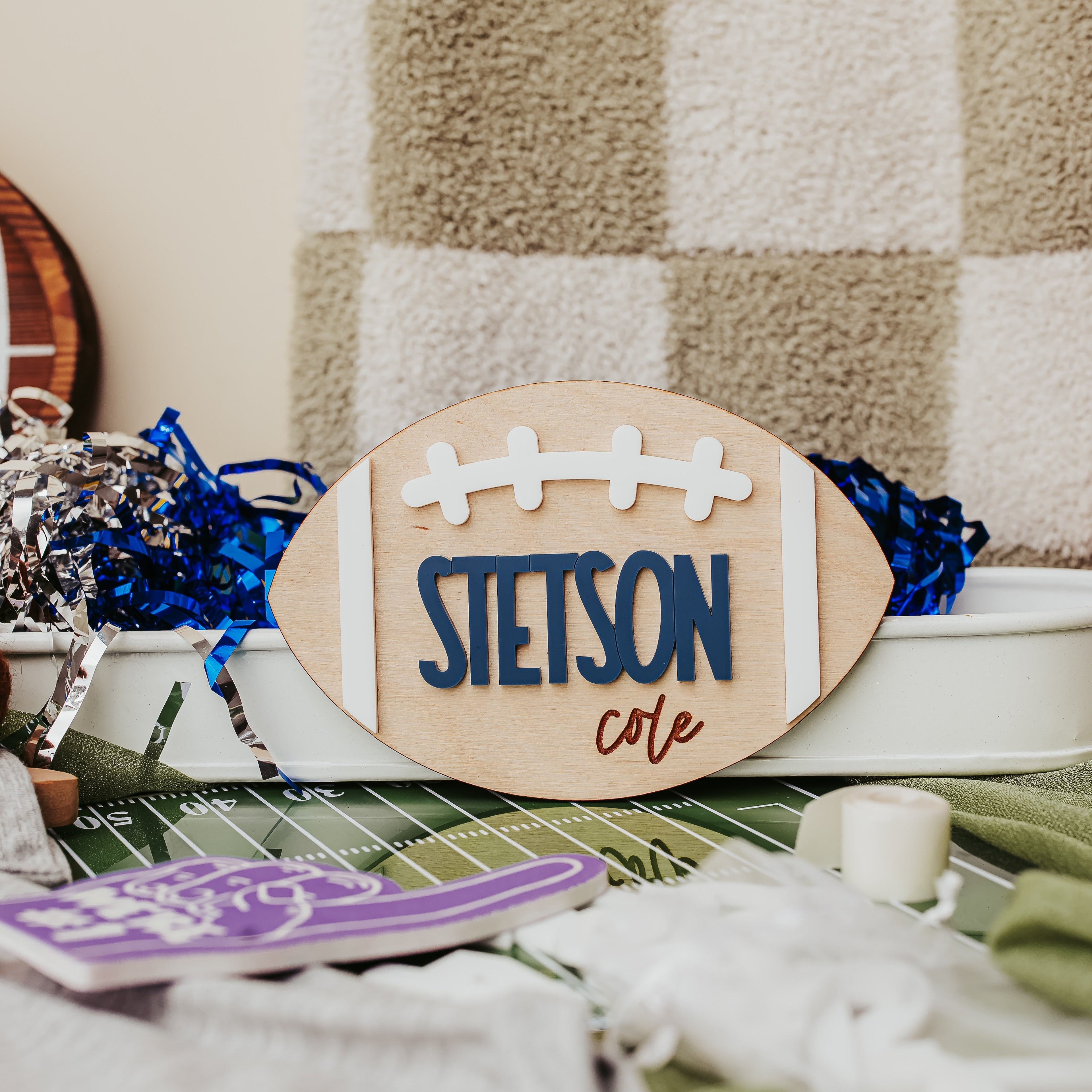 A custom wooden football-shaped sign reading 'STETSON cote' in navy and brown lettering, styled on a green turf mat with blue tinsel and a beige-and-beige checkered blanket in the background.
