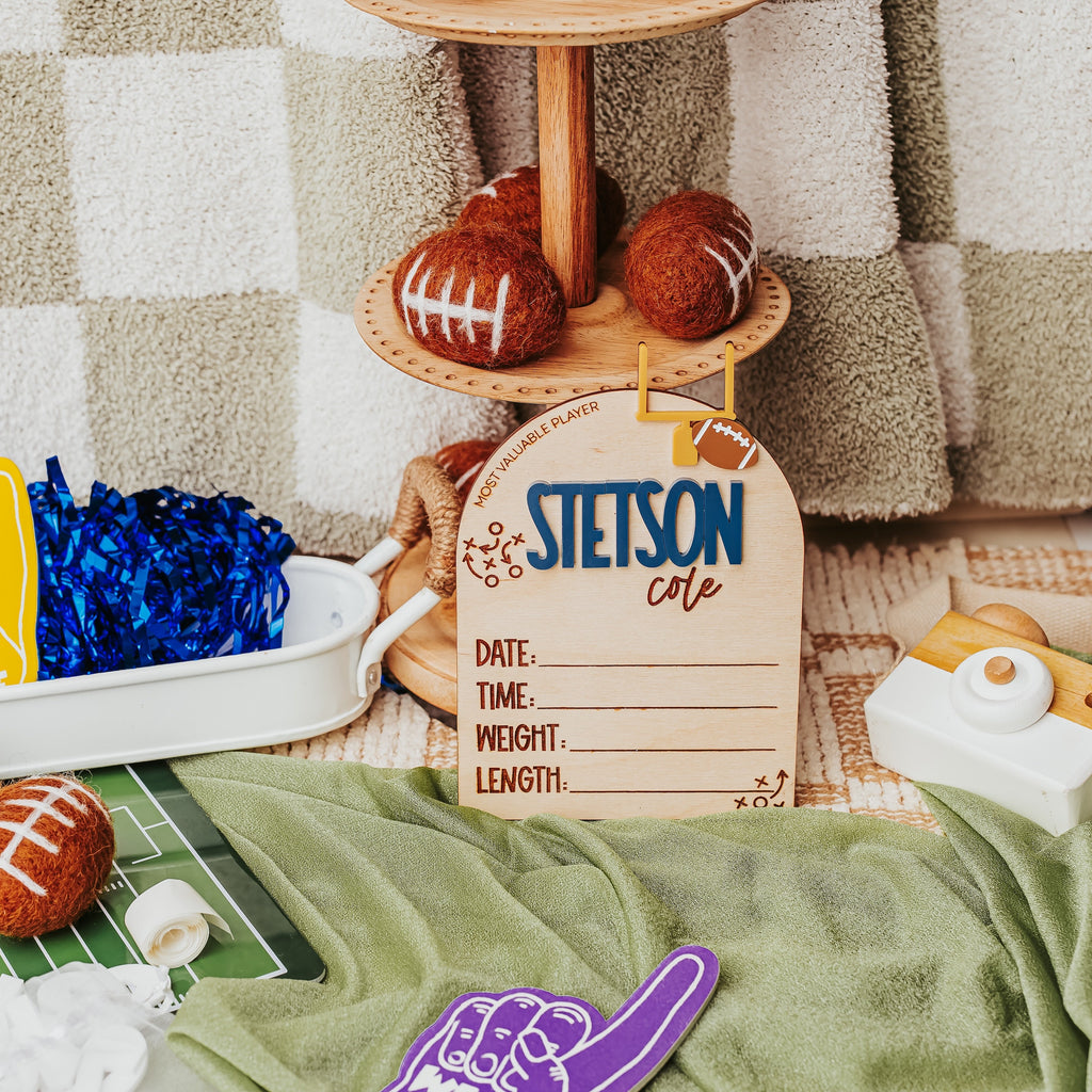 A styled football-themed baby birth stat  featuring wooden football birth stat signs, foam finger cutouts in yellow and purple, miniature footballs, a green field mat, and a checkered blanket backdrop.