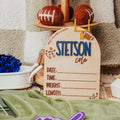 A wooden tiered stand holding football-themed props, including a wooden name sign that reads 'STETSON cole' with fields for date, time, weight, and length, set on a green blanket with a purple foam finger in the foreground and a checkered blanket behind.