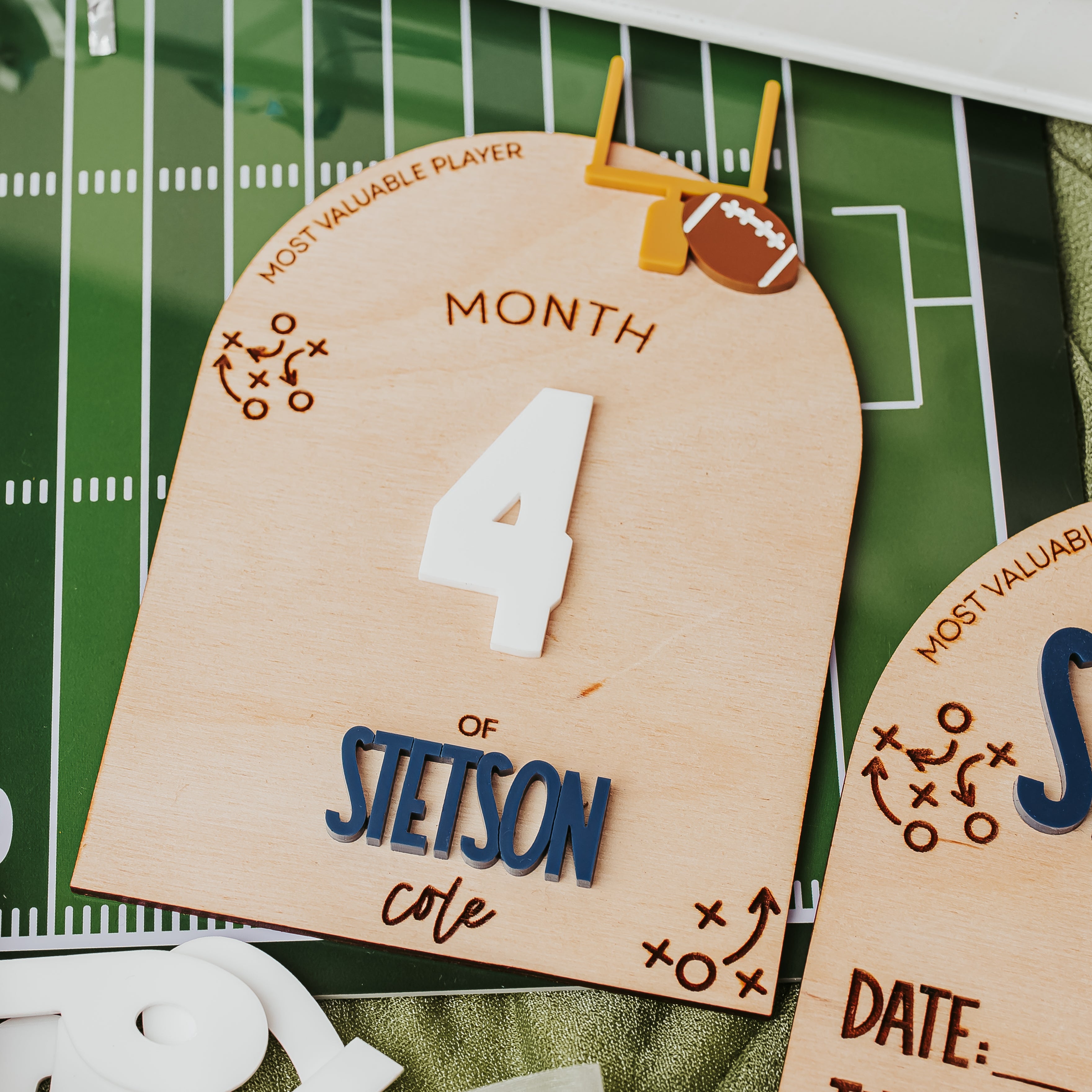 A close-up of a wooden football-themed monthly milestone card on a green football field mat, with part of another matching card visible at the right edge.