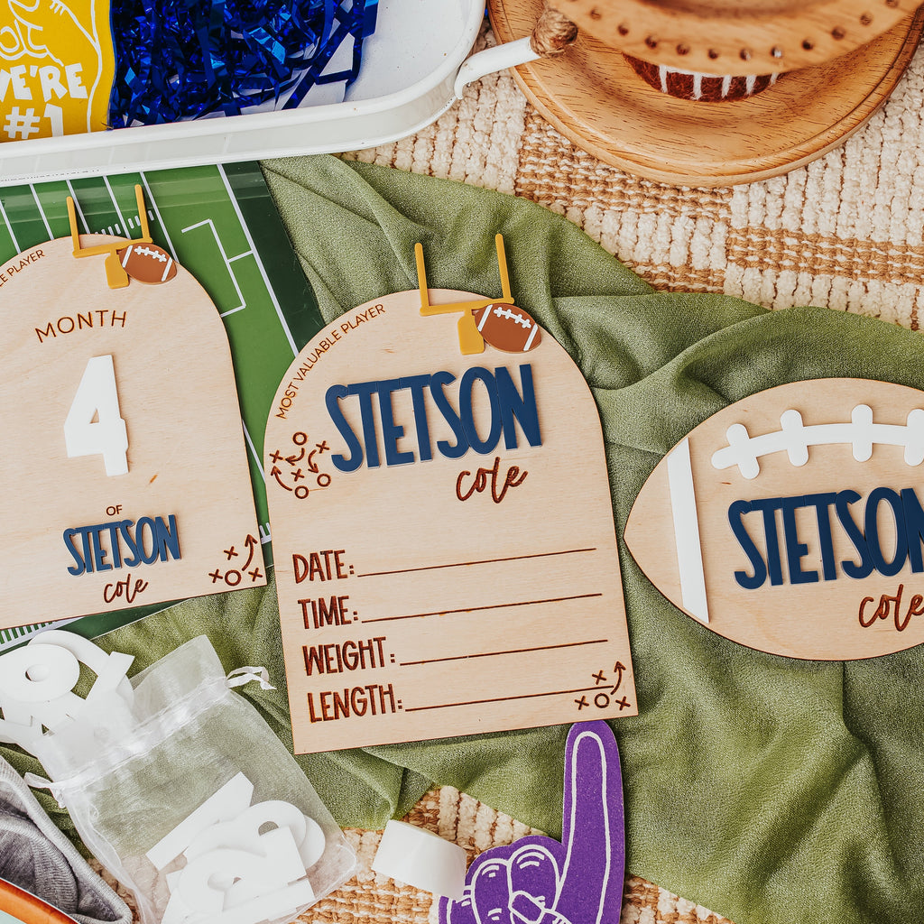Custom football-themed baby announcement laid out on a green blanket, including three wooden name tags with football graphics, a purple foam finger, and other party accessories.