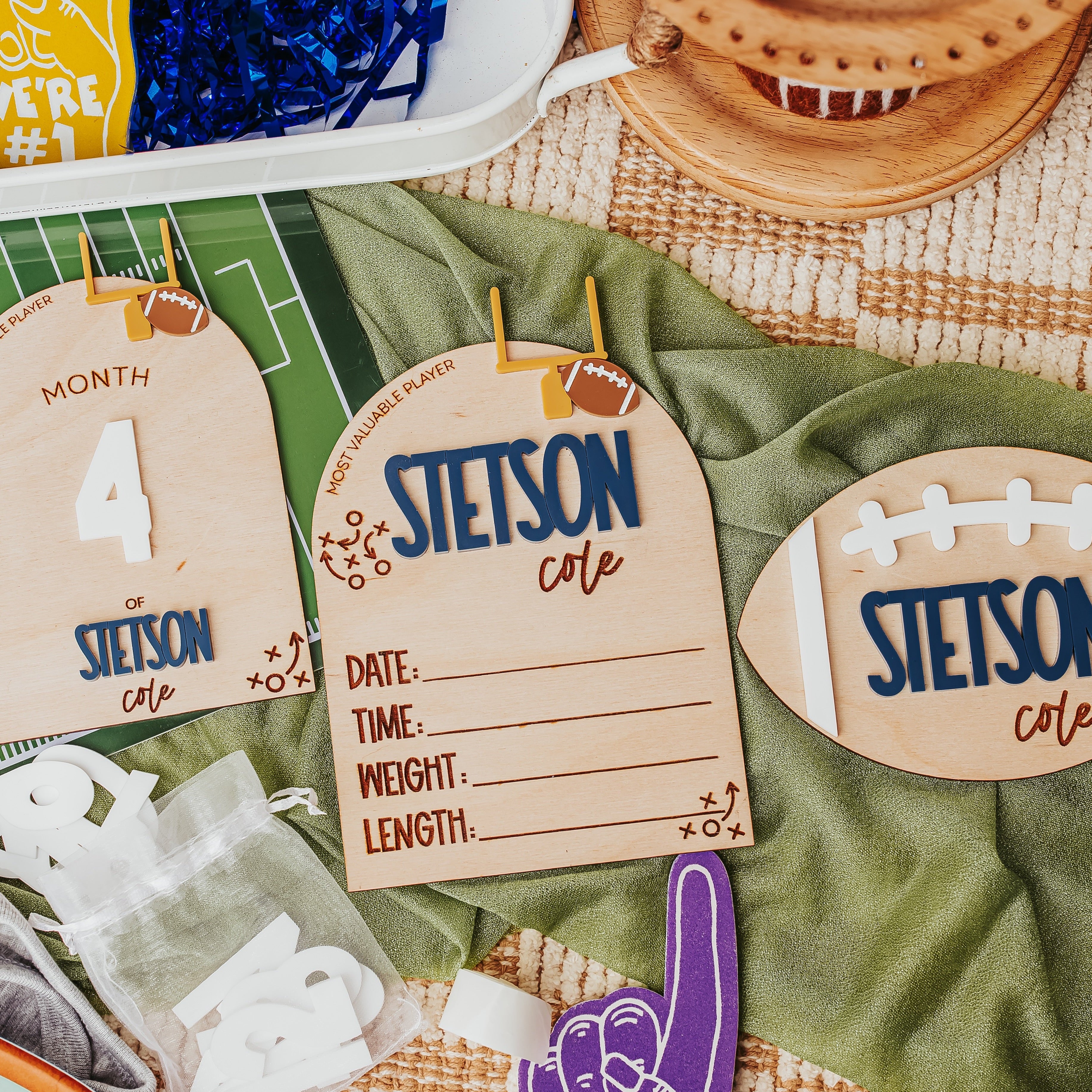Custom football-themed baby announcement laid out on a green blanket, including three wooden name tags with football graphics, a purple foam finger, and other party accessories.