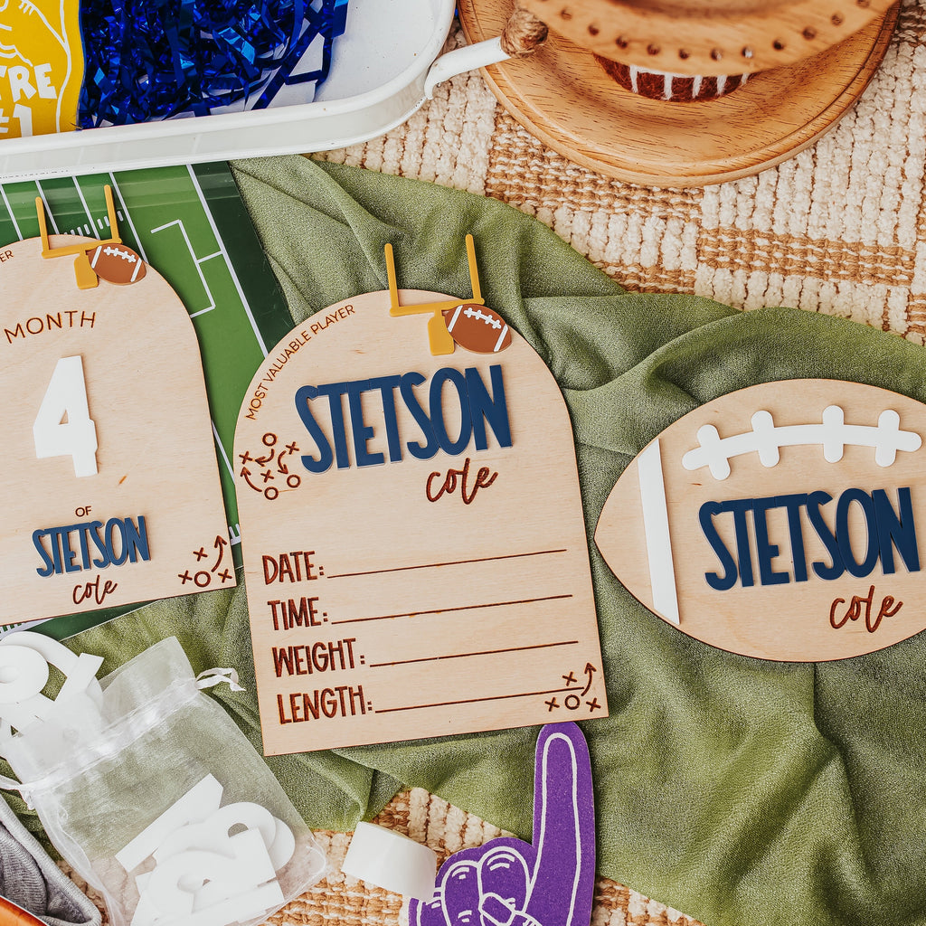 Custom football-themed baby milestone cards laid out on a green blanket with sports props, including a football-shaped card, a month card, and a birth details card.